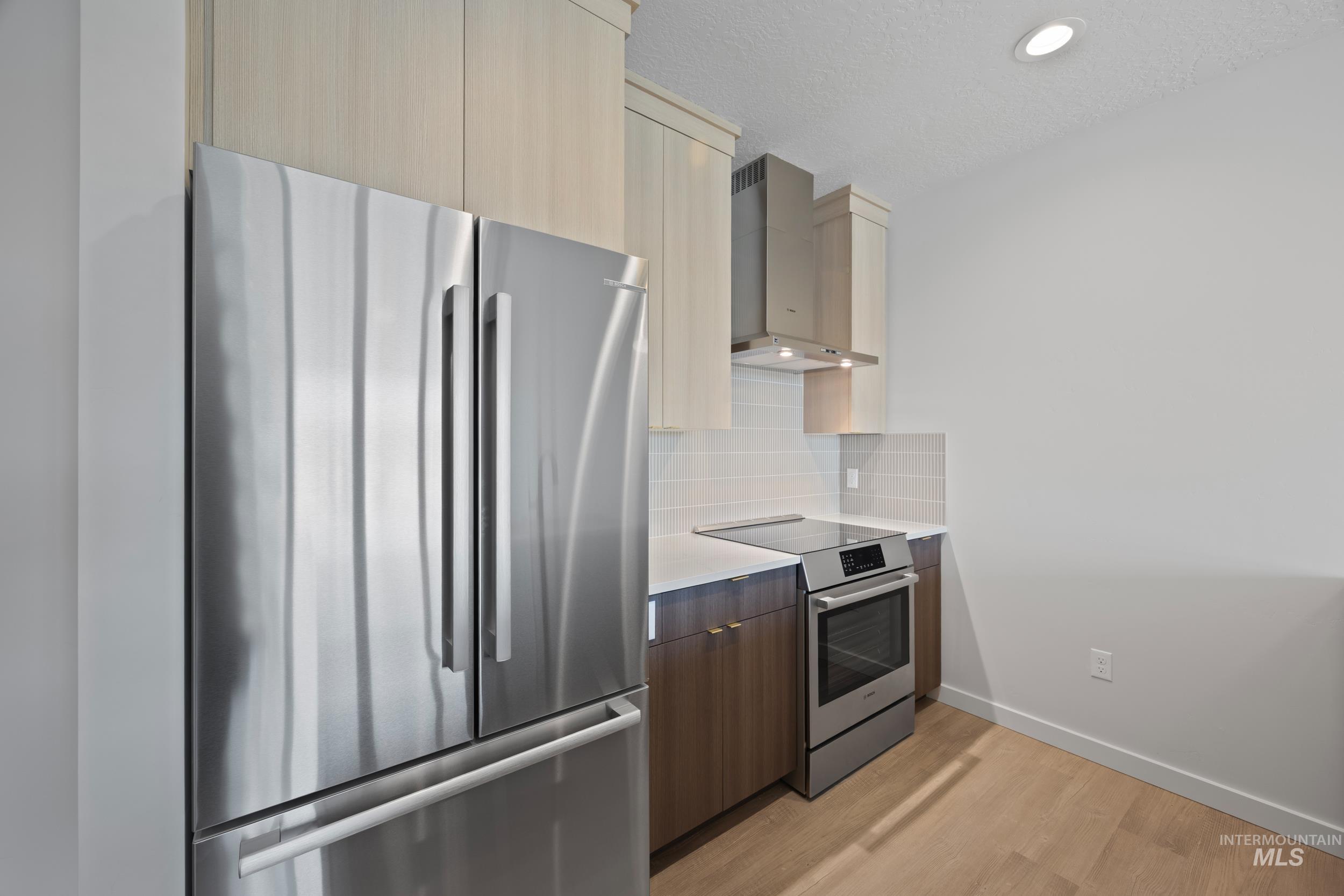 Kitchen with modern cabinets, appliances with stainless steel finishes, wall chimney range hood, light wood-type flooring, and decorative backsplash