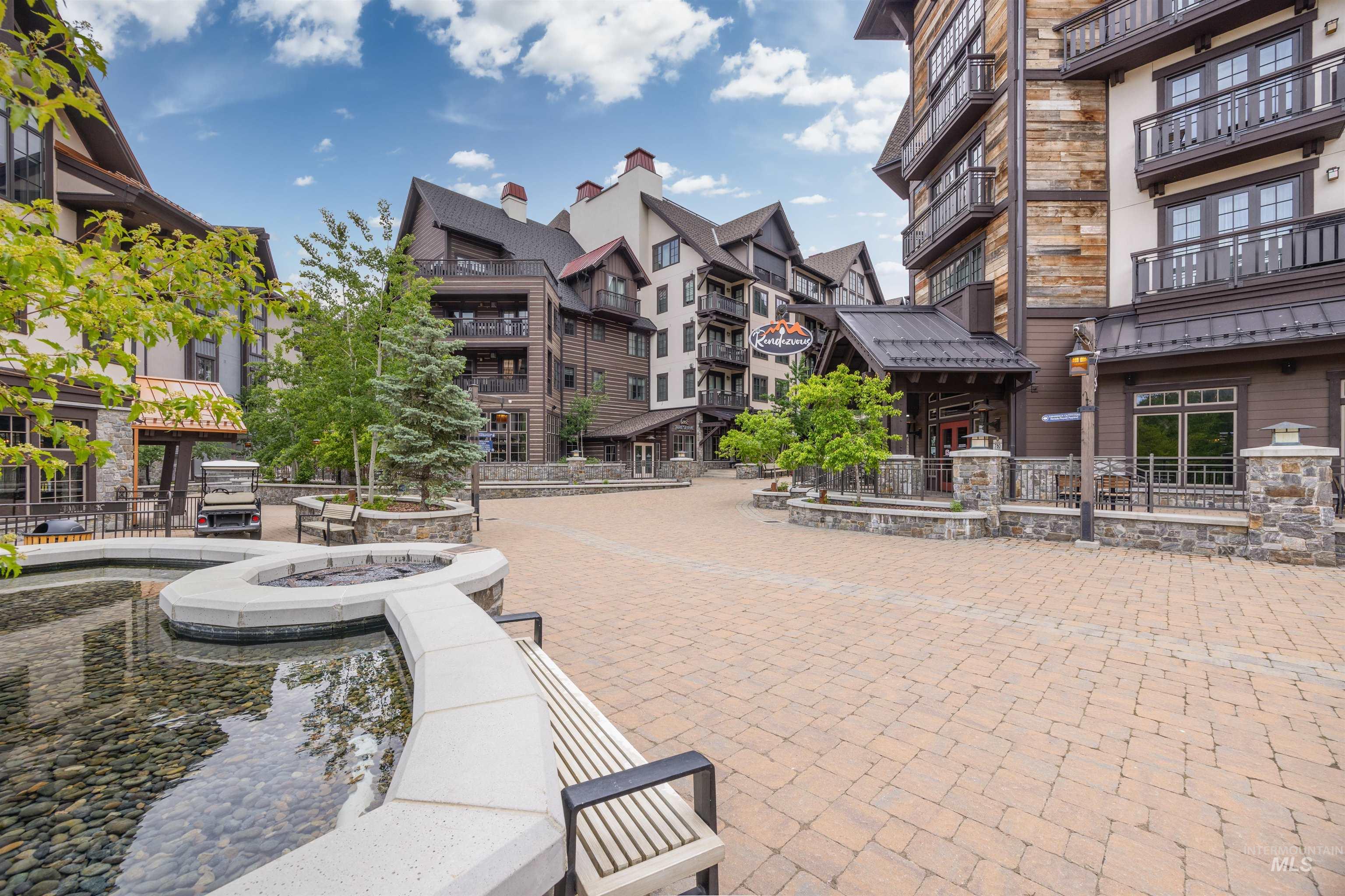 Expansive paver patio featuring a stone-lined water feature, multiple seating areas, mature landscaping, and architectural buildings with balconies and varied exterior finishes