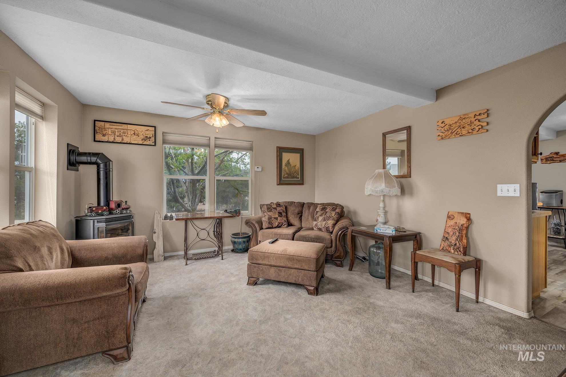 Carpeted living room with a wood stove, arched walkways, a ceiling fan, beam ceiling, and a textured ceiling