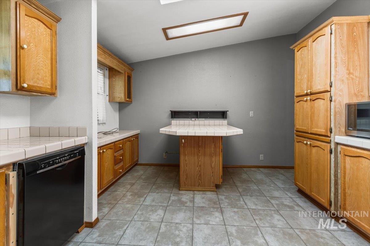 Kitchen featuring black dishwasher, tile countertops, lofted ceiling, brown cabinetry, and light tile patterned flooring