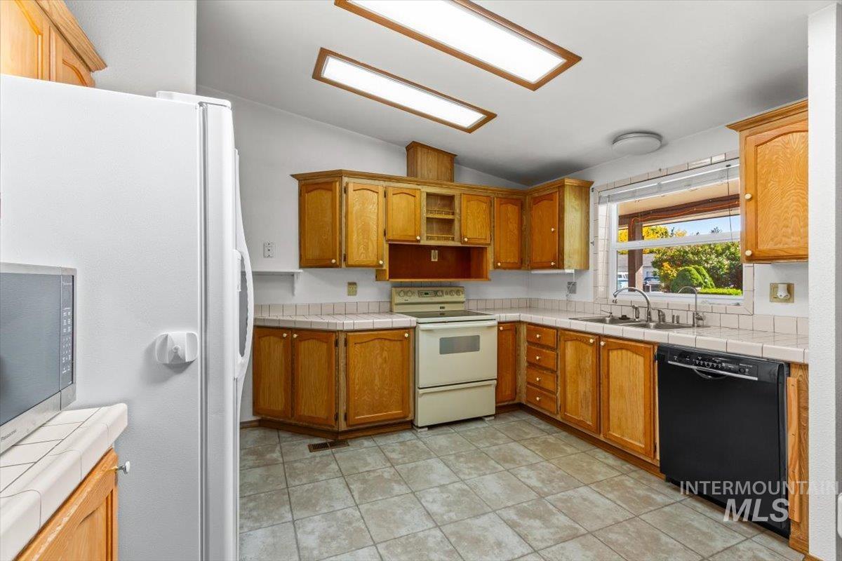 Kitchen featuring tile counters, vaulted ceiling, white appliances, brown cabinetry, and light tile patterned flooring