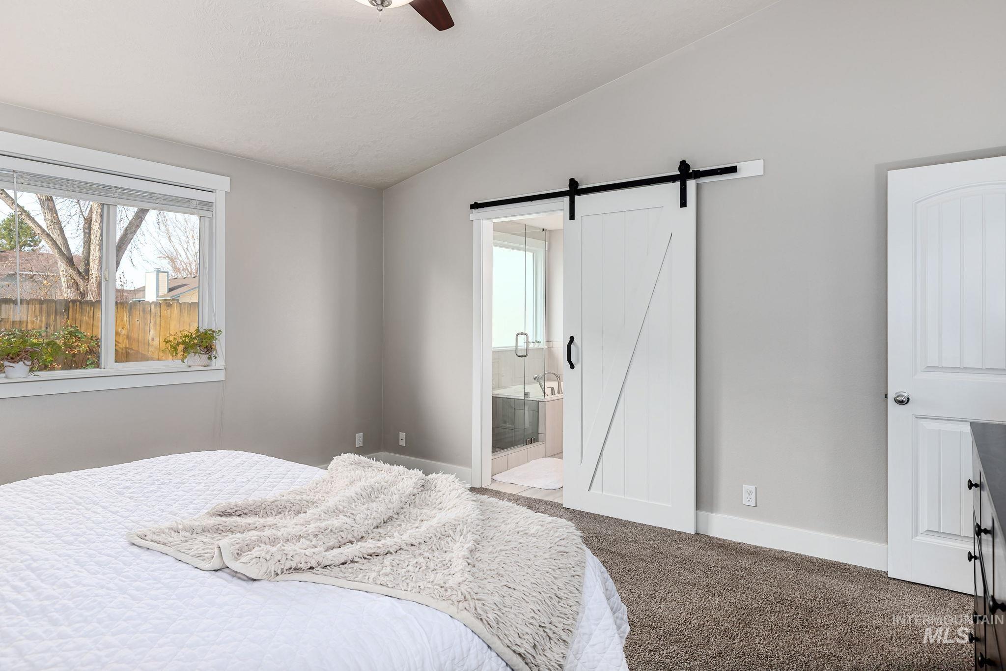 Carpeted bedroom with vaulted ceiling, a barn door, ensuite bathroom, and a ceiling fan
