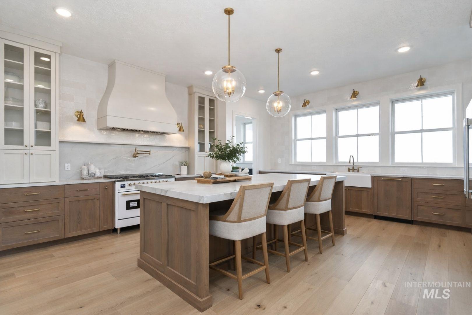 Gourmet kitchen featuring a large central island with seating, a white apron-front sink, light wood-finish flooring, a decorative range hood, and multiple windows