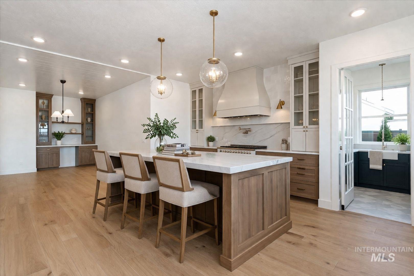 Spacious kitchen featuring a large central island with a white countertop, light wood-finish flooring, recessed lighting, a built-in range hood, and glass-paneled double doors leading to an adjacent room with a window and sink