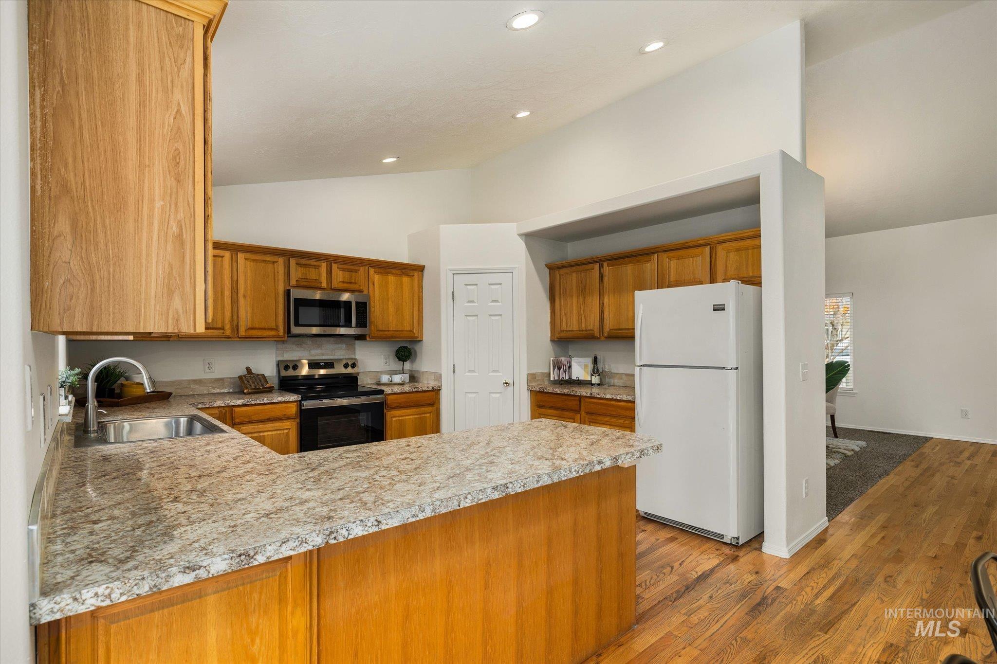 Kitchen with vaulted ceiling, stainless steel appliances, brown cabinetry, light countertops, and a peninsula