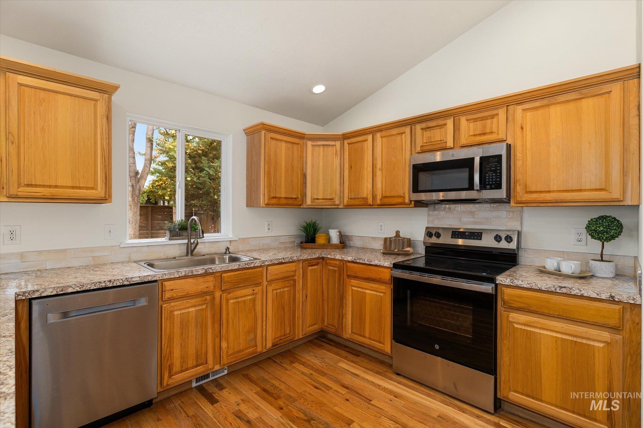 Kitchen with stainless steel appliances, light wood-style floors, brown cabinetry, light countertops, and lofted ceiling