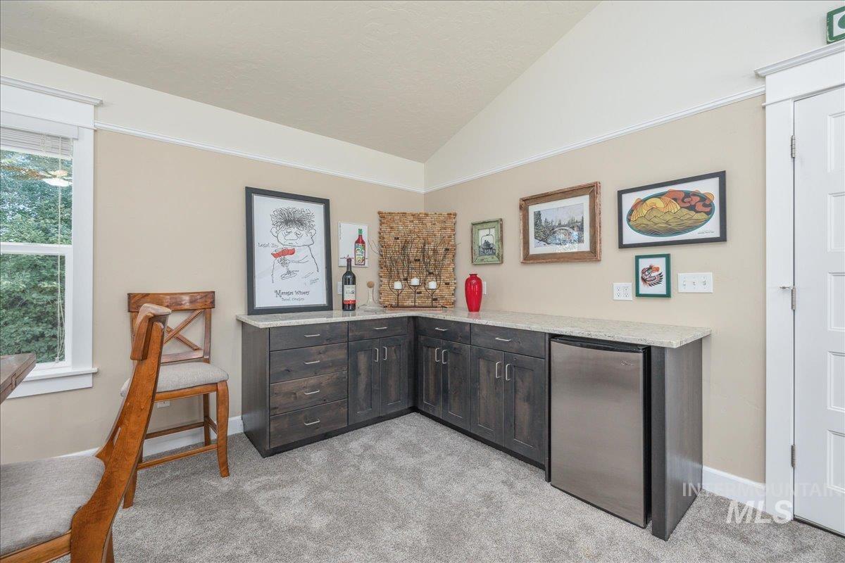 Bar featuring vaulted ceiling, light colored carpet, light stone counters, stainless steel fridge, and dark brown cabinets