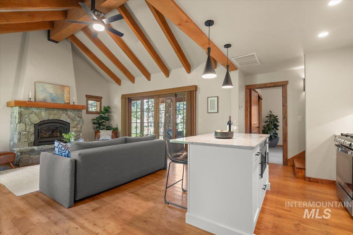 Vaulted ceilings with exposed wood beams, stone fireplace with wood mantel, wood-finish flooring, white kitchen island with a light-toned countertop, and modern pendant lighting