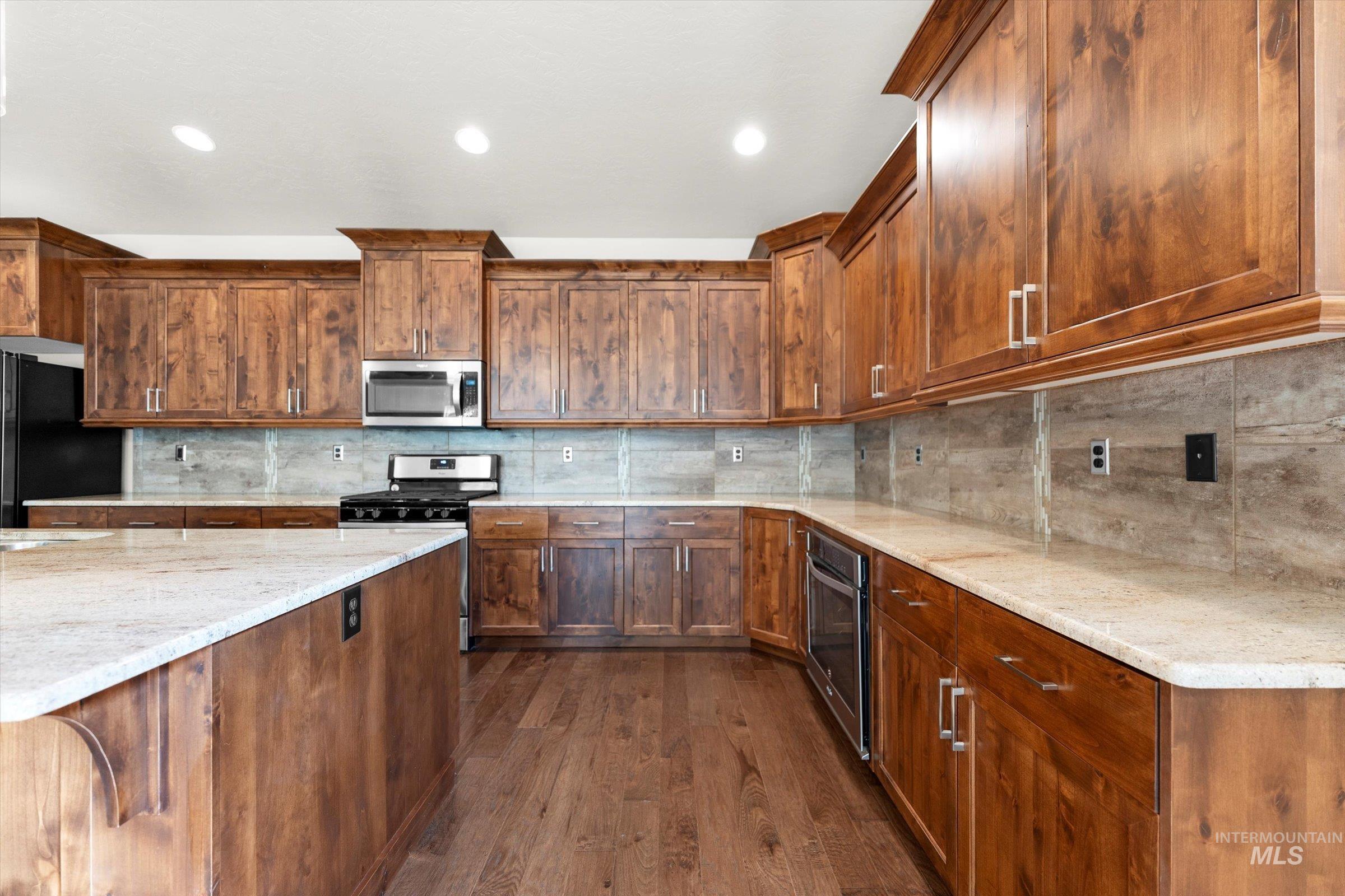 Kitchen featuring backsplash, dark wood-style floors, appliances with stainless steel finishes, light stone countertops, and brown cabinets
