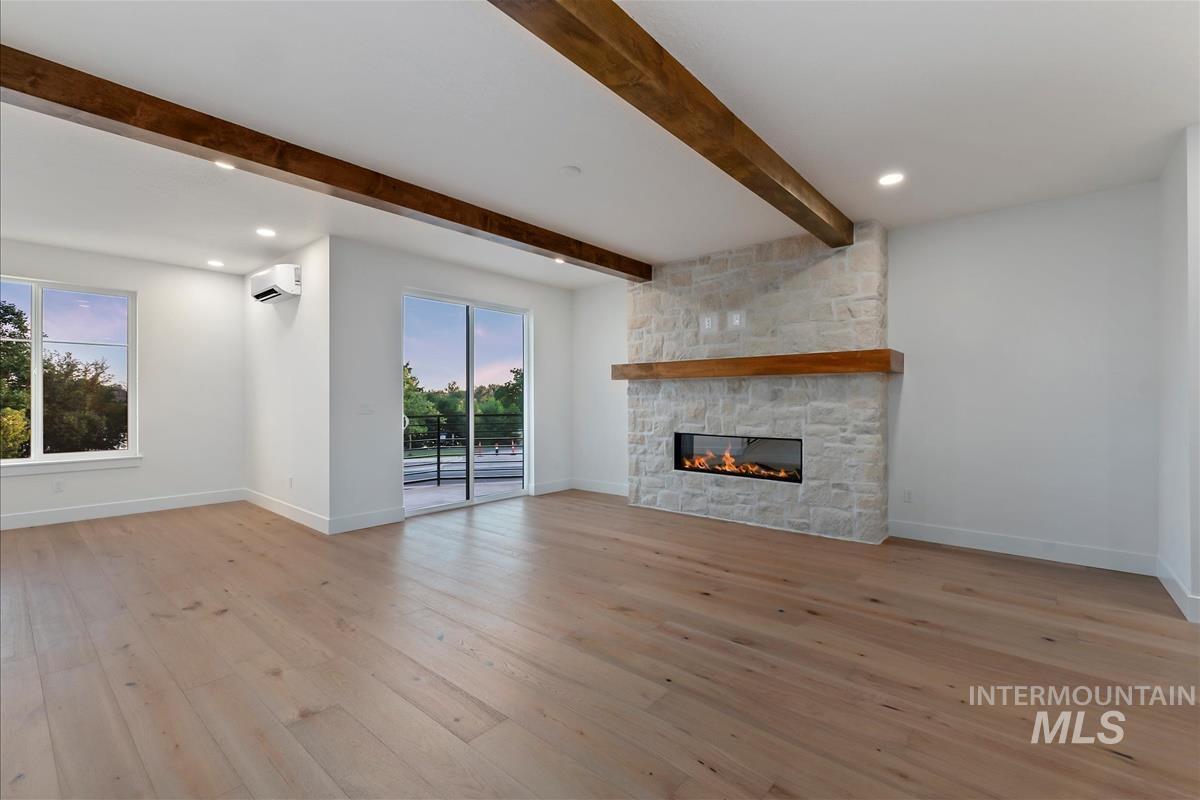 Expansive living space featuring light wood-finish flooring, a stone-clad fireplace with a wood mantel, exposed ceiling beams, recessed lighting, and a sliding glass door to an exterior balcony