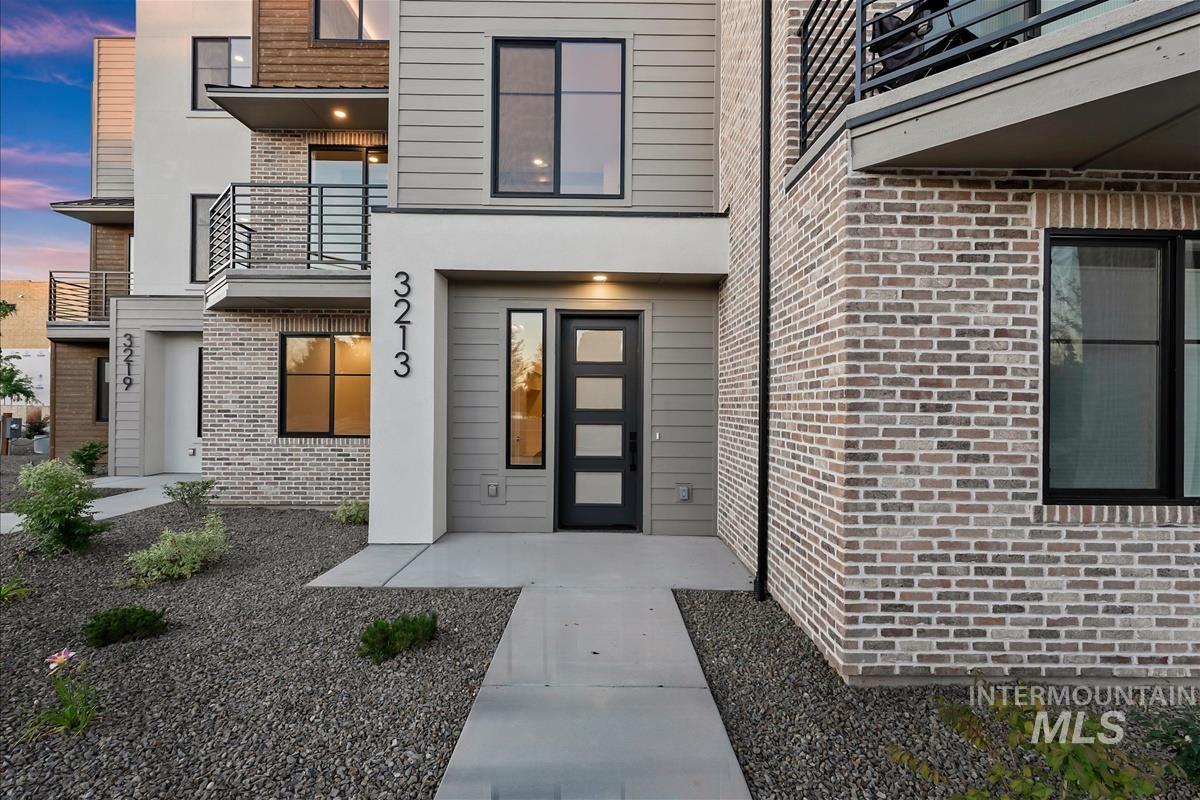Contemporary multi-level residence featuring a modern dark-paneled entry door, contrasting light brick and horizontal siding, black-framed windows, and an upper-level balcony with metal railings