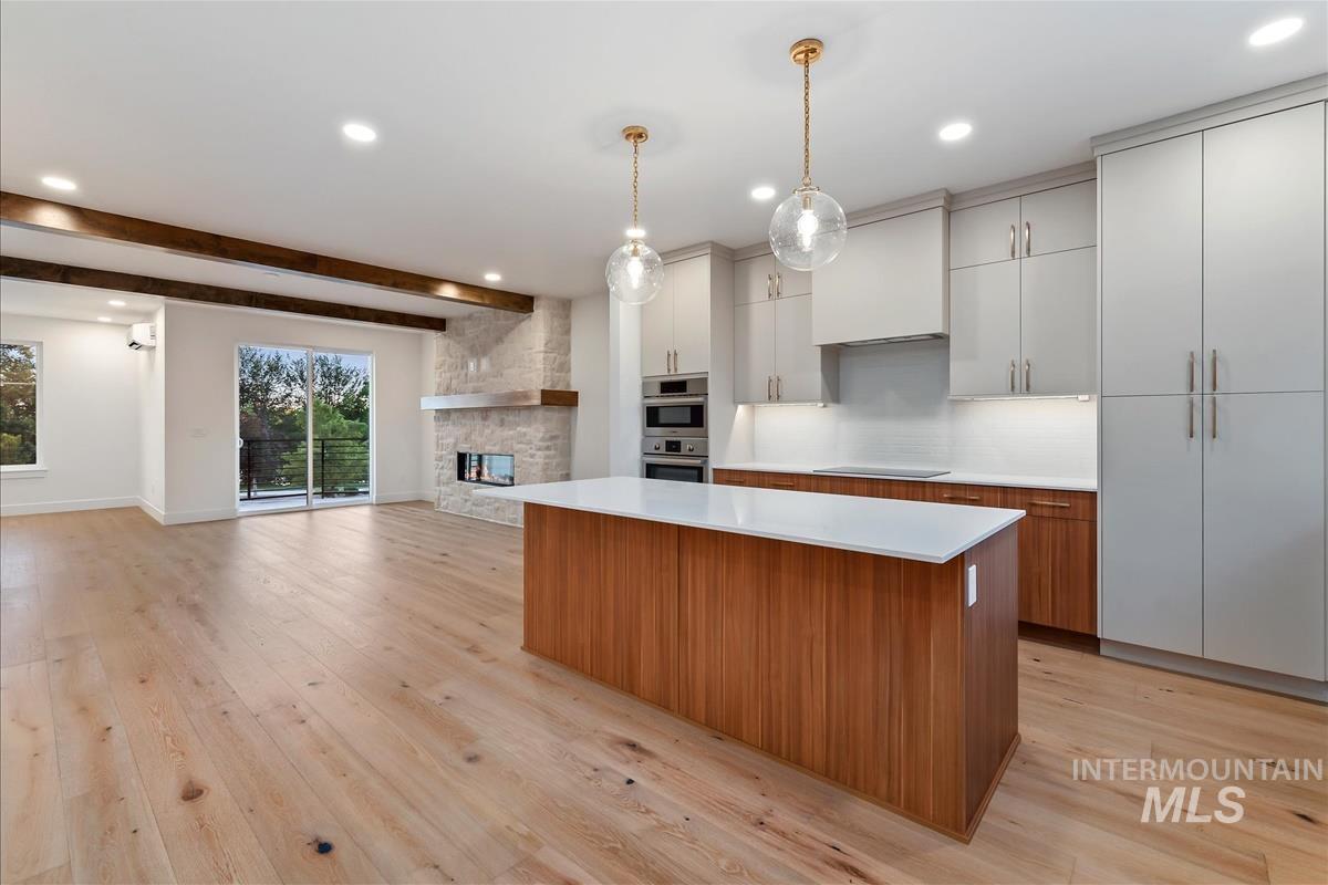 Open-concept living space featuring wide plank wood-finish flooring, a contemporary kitchen island with a white countertop, light-toned cabinetry, and a stone-clad fireplace with a natural wood mantel