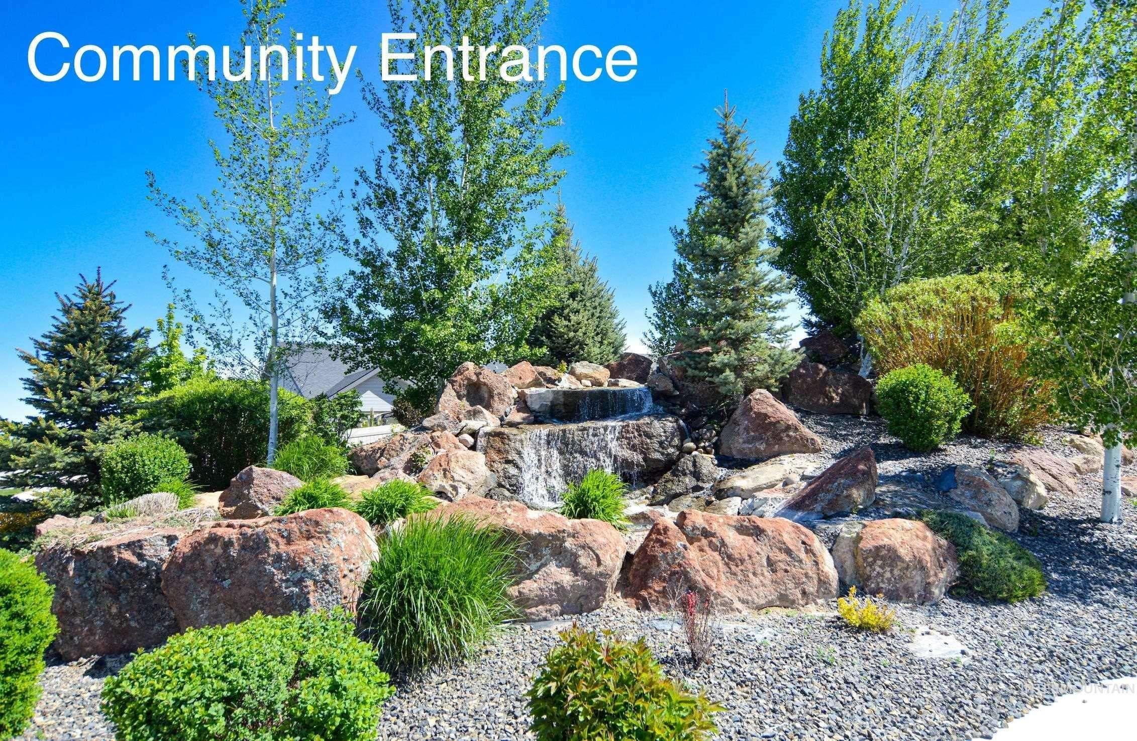 Landscaped community entrance featuring a rock waterfall, mature trees, diverse shrubs, and decorative gravel