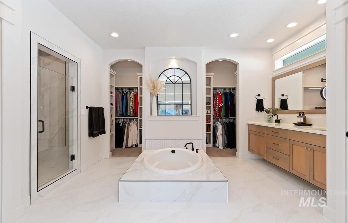 Spacious bathroom featuring a built-in soaking tub with marble-look surround, a glass-enclosed shower, dual vanity with light wood cabinetry and stone countertops, and two arched alcoves with built-in shelving