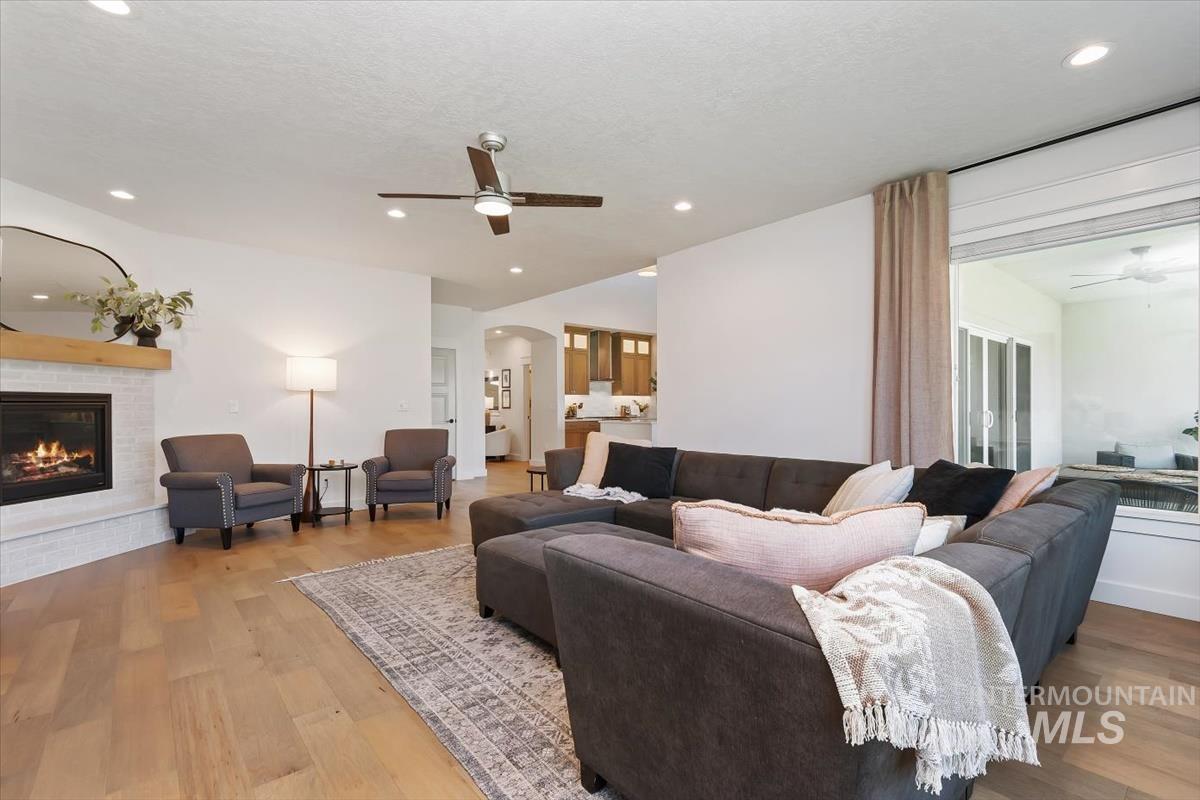 Spacious living area featuring light wood-finish flooring, a white brick fireplace with a wood mantel, a ceiling fan with integrated lighting, recessed lighting, and a large sliding glass door