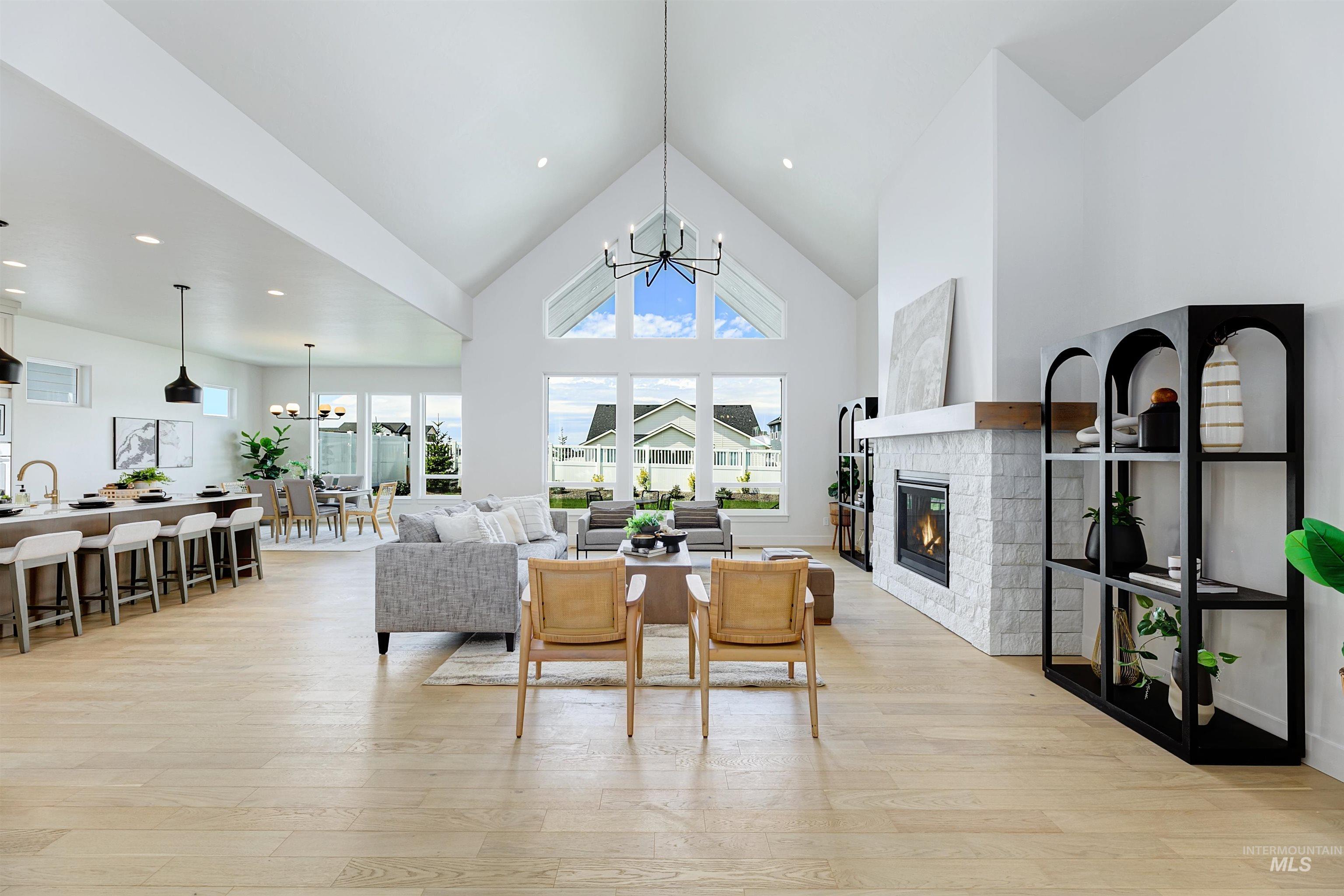 Expansive great room featuring vaulted ceilings with a linear chandelier, light wood-finish flooring, a contemporary fireplace with a stone surround and wood mantel, and large windows providing abundant natural light