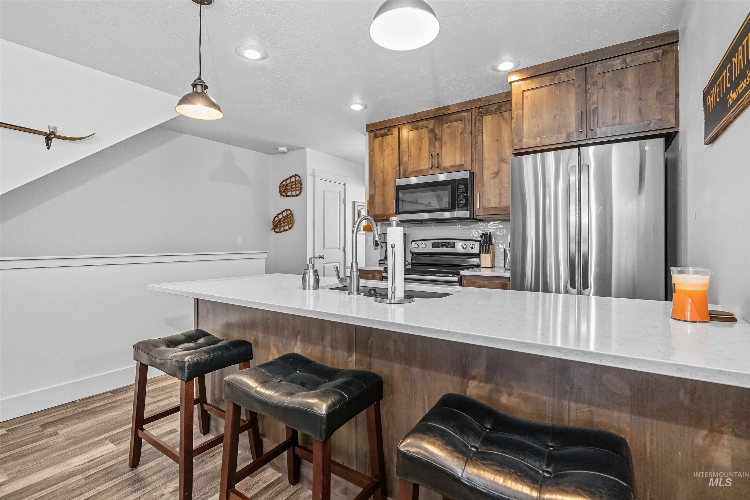 Kitchen with appliances with stainless steel finishes, hanging light fixtures, light stone counters, light wood finished floors, and brown cabinetry