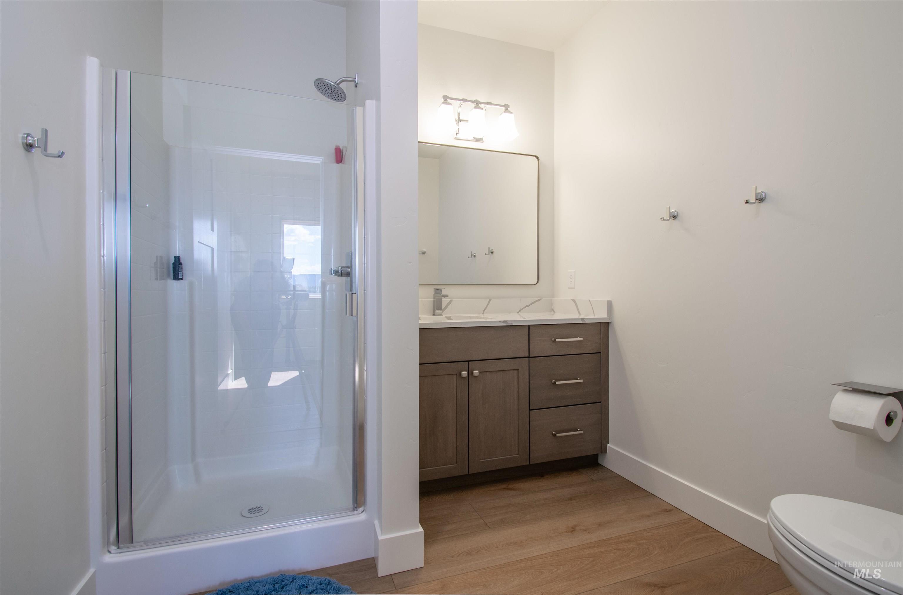 Bathroom featuring a glass-enclosed shower, single vanity with a light gray countertop, wood-finish cabinetry, a rectangular mirror, and wood-finish flooring