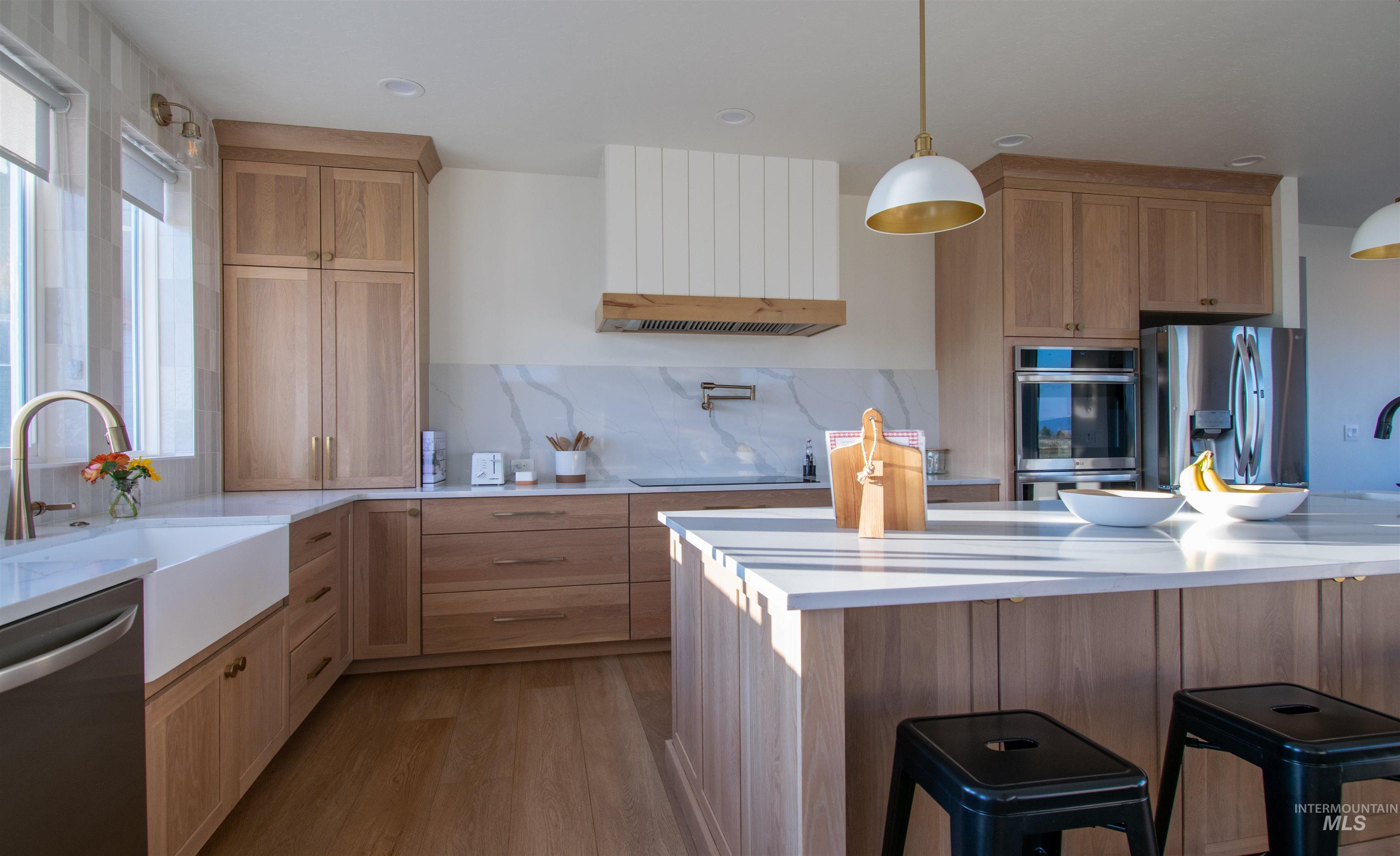 Gourmet kitchen featuring a large central island, white apron-front sink, light wood-finish cabinetry, stainless steel appliances, and a marble-finish backsplash