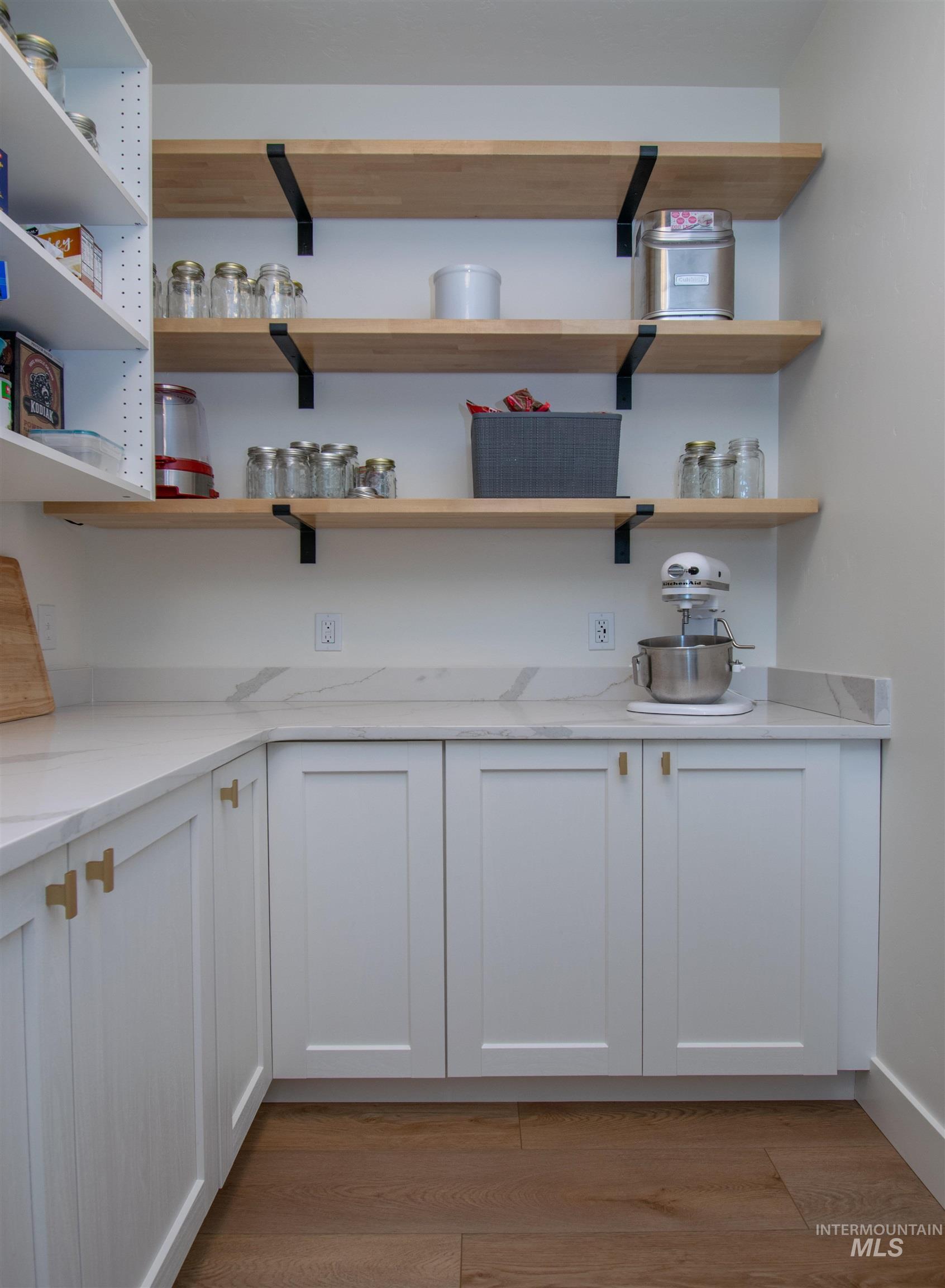 Spacious pantry featuring white shaker-style cabinetry with gold-tone hardware, a white countertop with subtle veining, and open wood shelving with matte black brackets