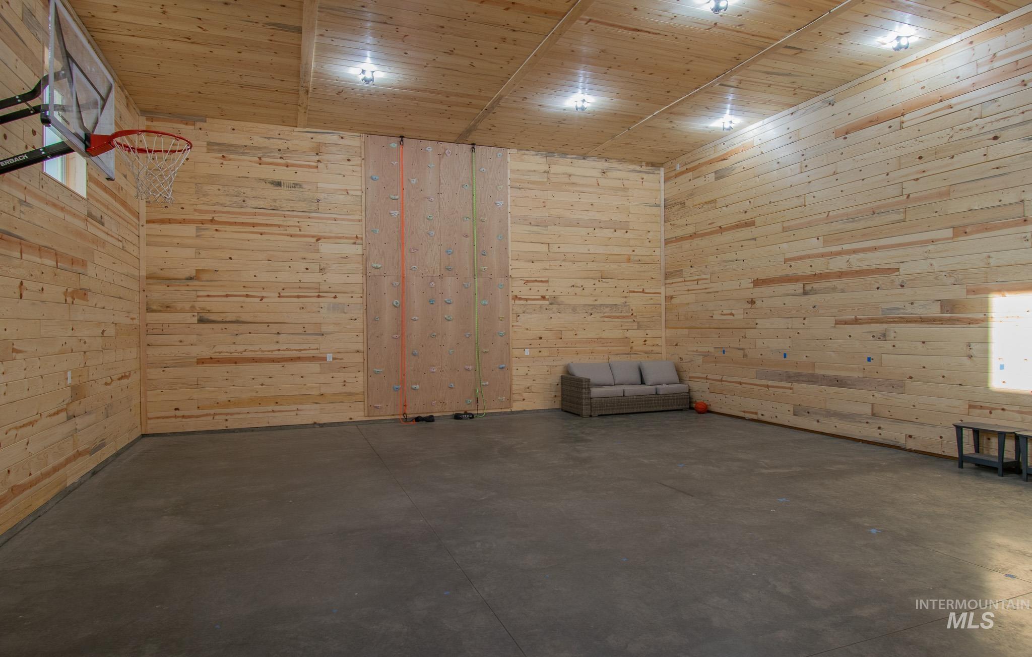 Spacious indoor recreational area featuring wood-paneled walls and ceiling, polished concrete flooring, a built-in climbing wall, a basketball hoop, and recessed lighting fixtures