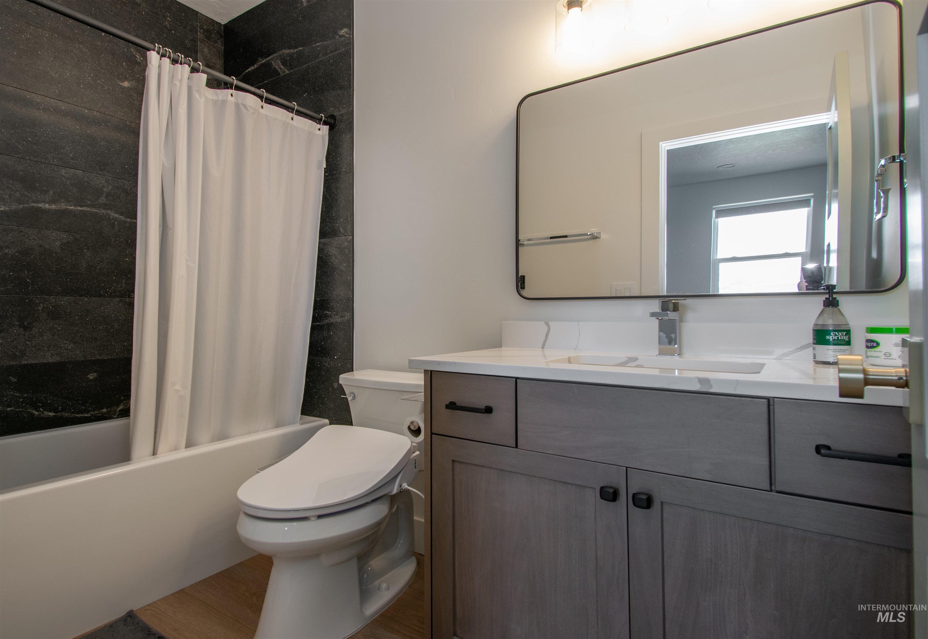 Bathroom featuring a grey wood-finish vanity with a white countertop and integrated sink, a rectangular mirror, a bathtub with dark stone-look tile surround, and light wood-finish flooring