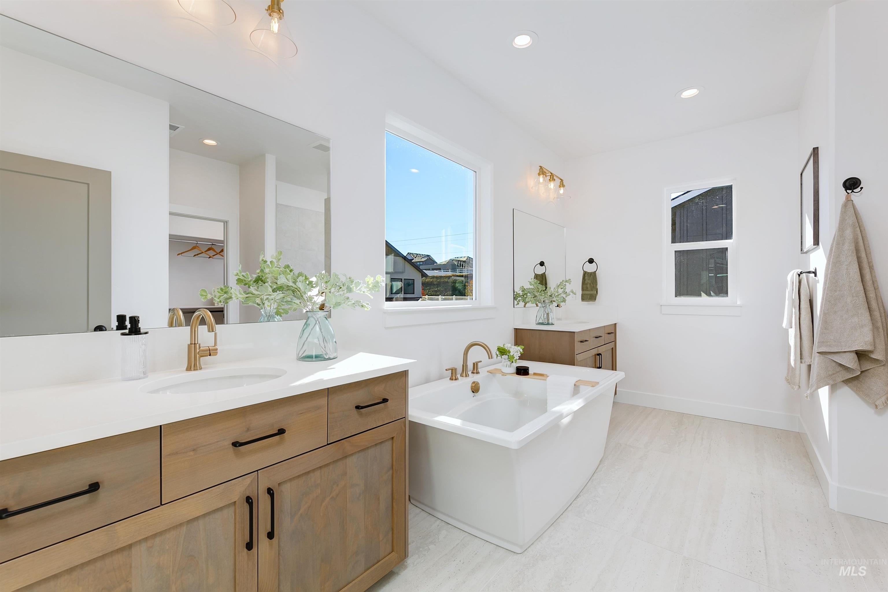 Bright bathroom featuring a freestanding soaking tub, dual wood-finish vanities with solid surface countertops, light-toned tile flooring, and recessed ceiling lighting