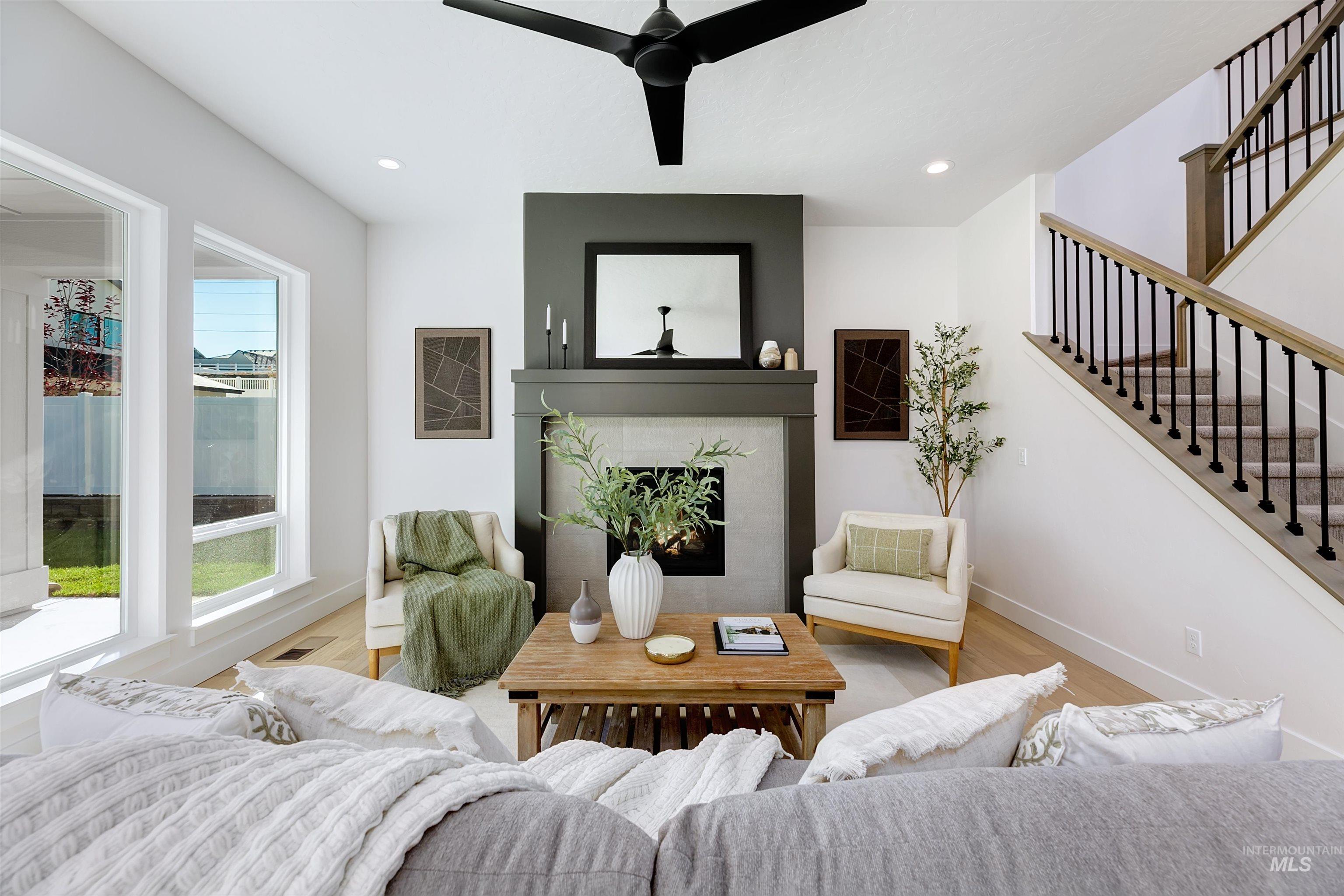 Living area featuring a floor-to-ceiling fireplace with a dark accent mantel, a modern black ceiling fan, light wood-finish flooring, and a carpeted staircase with an open spindle railing