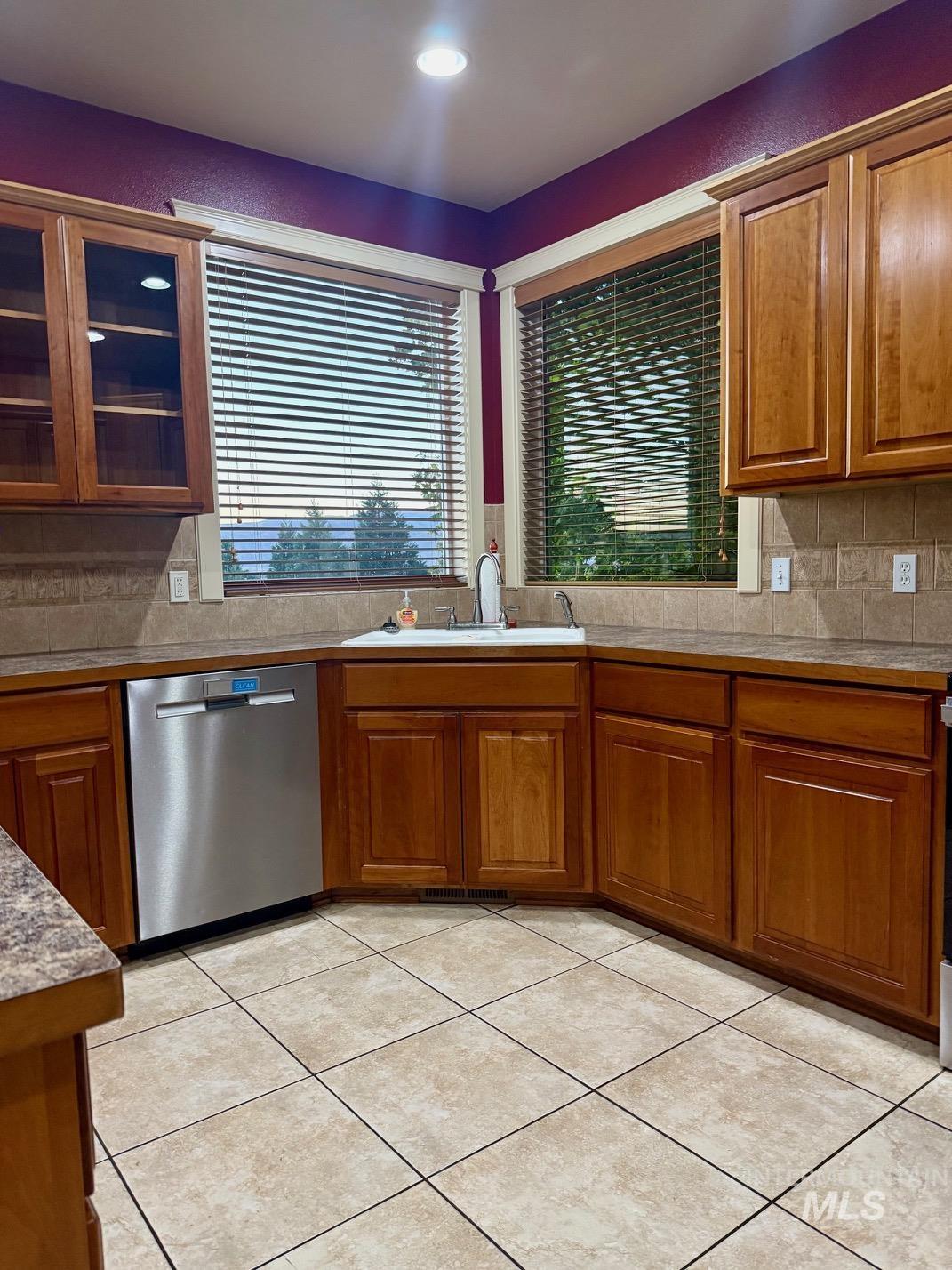 Kitchen with wood finish cabinetry, decorative backsplash, dishwasher, glass insert cabinets, and light tile patterned floors