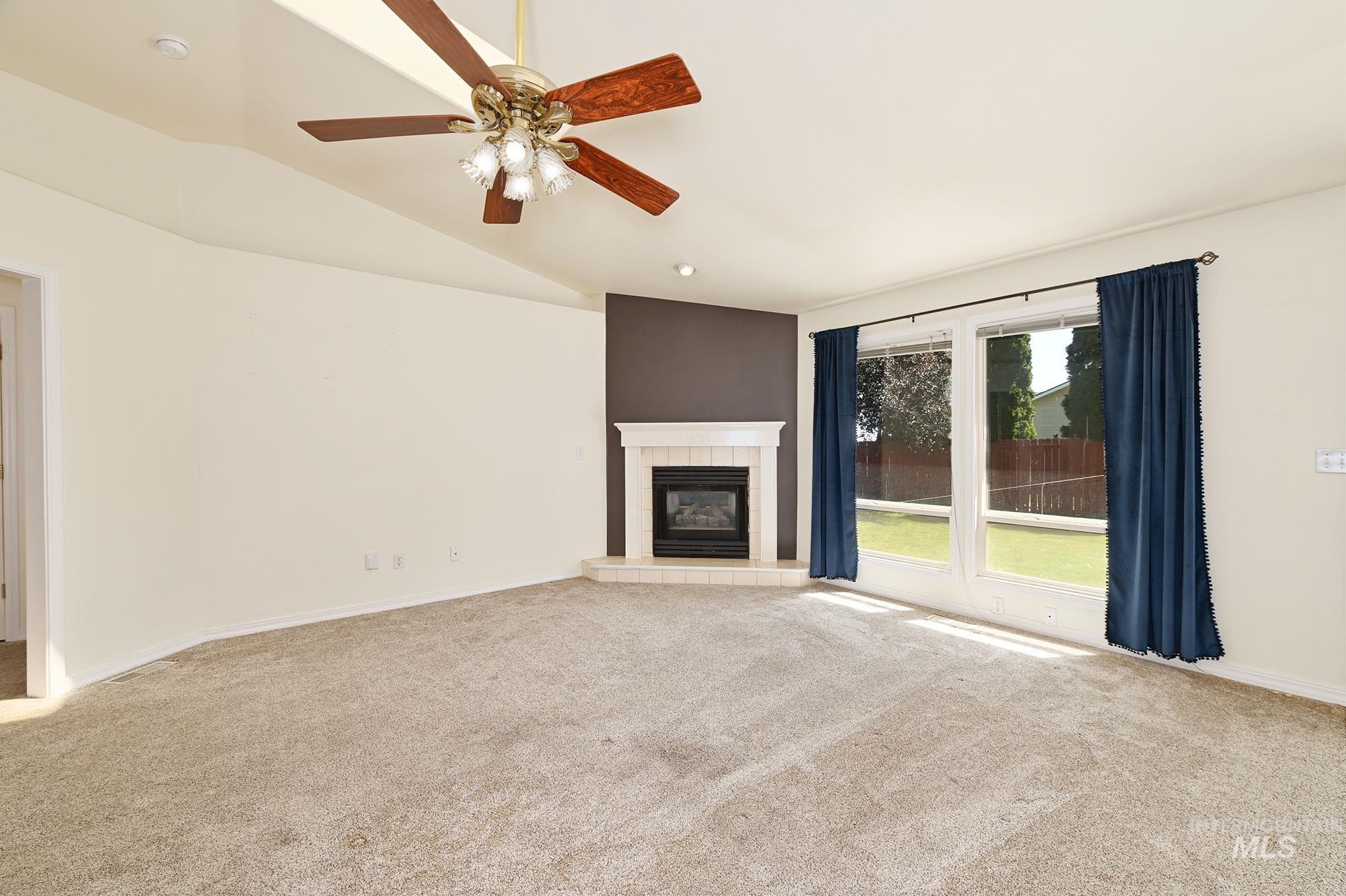 Spacious interior room featuring a corner fireplace with a white mantel and hearth, large windows overlooking a green lawn, and a ceiling fan with integrated lighting