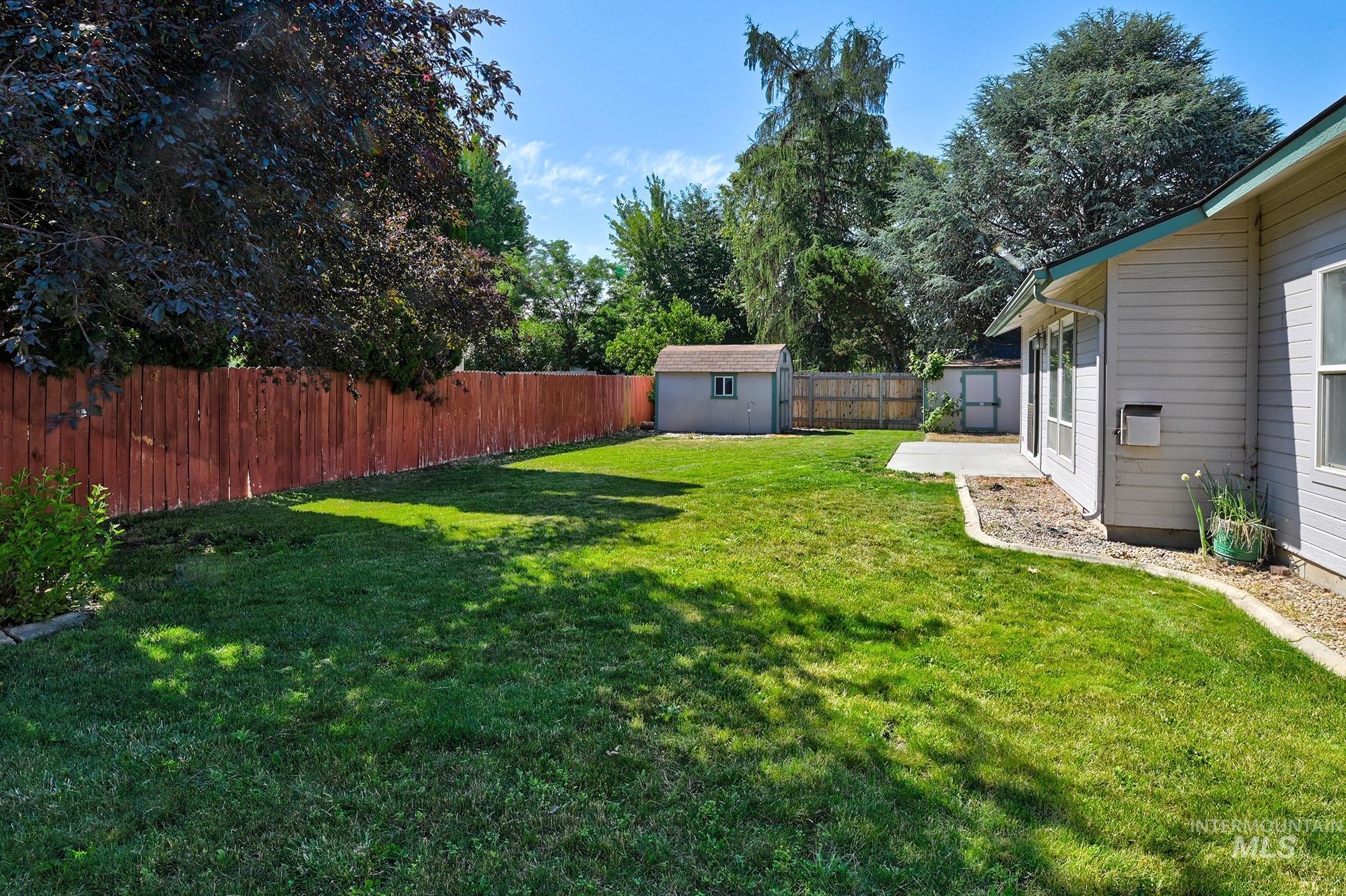 Expansive backyard featuring a manicured lawn, full privacy fencing, established shade trees, a concrete patio, and a dedicated storage shed