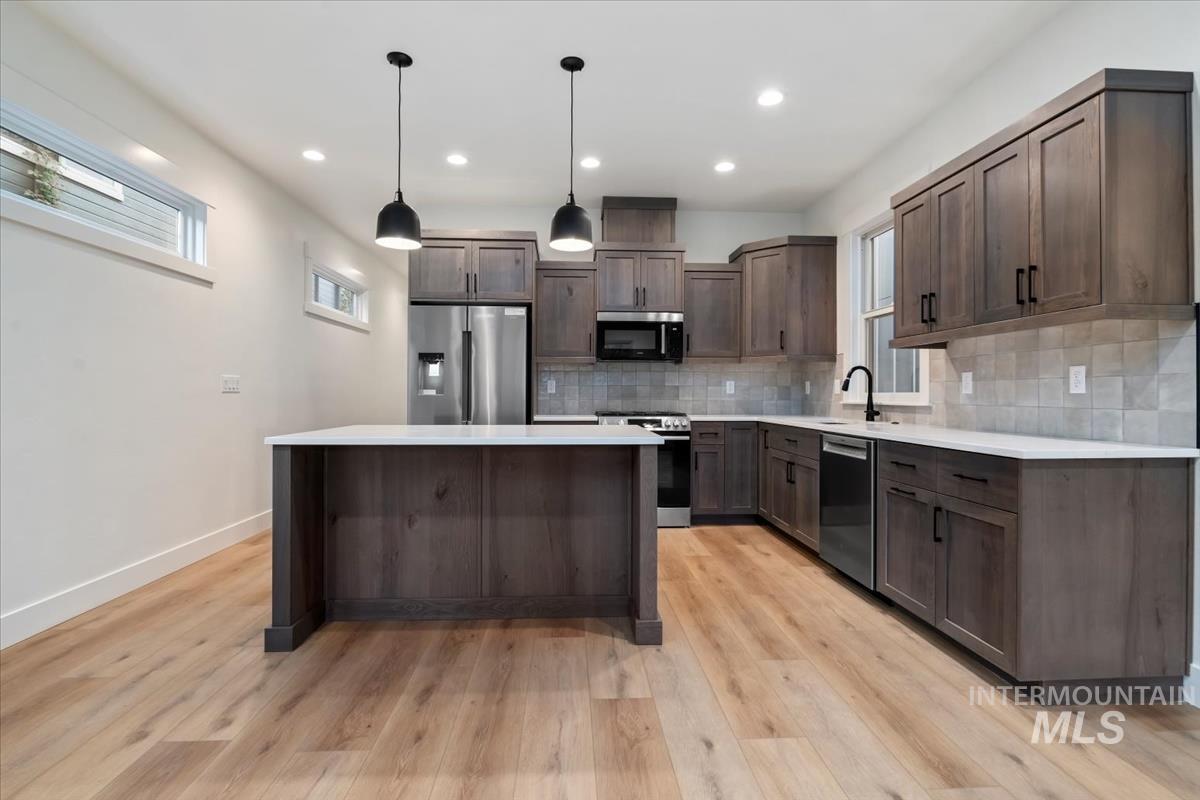 Kitchen featuring appliances with stainless steel finishes, tasteful backsplash, dark brown cabinetry, pendant lighting, and light wood-type flooring
