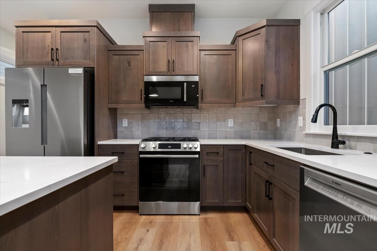 Kitchen with stainless steel appliances, decorative backsplash, light wood-style floors, dark brown cabinetry, and light stone counters