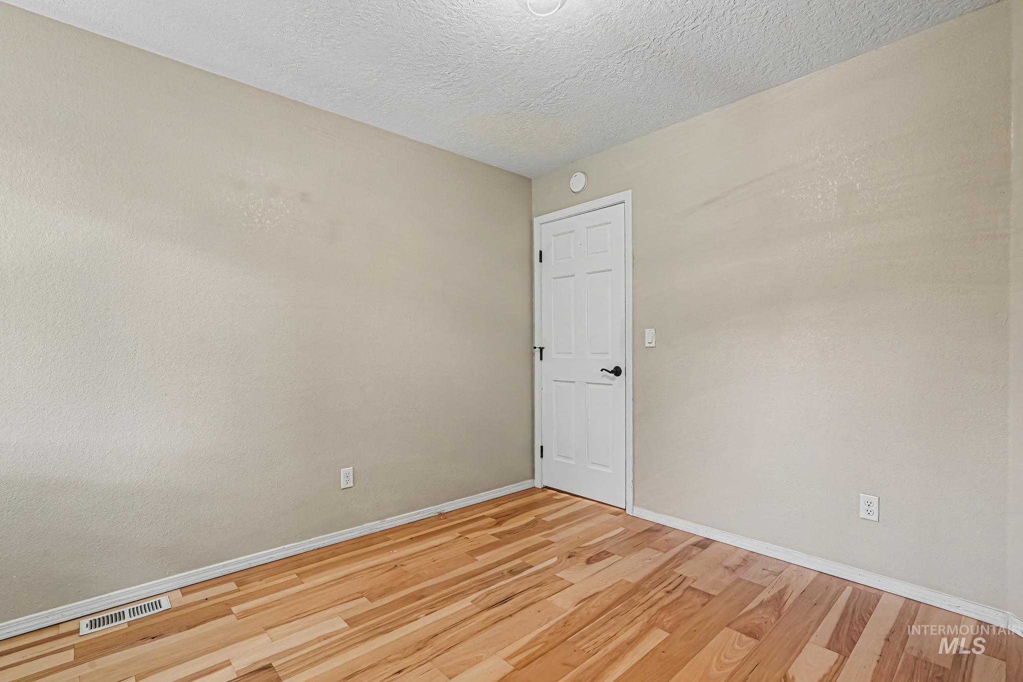 Interior room featuring light-toned walls, wood-finish flooring, a white paneled door with black hardware, and an overhead flush-mount light fixture