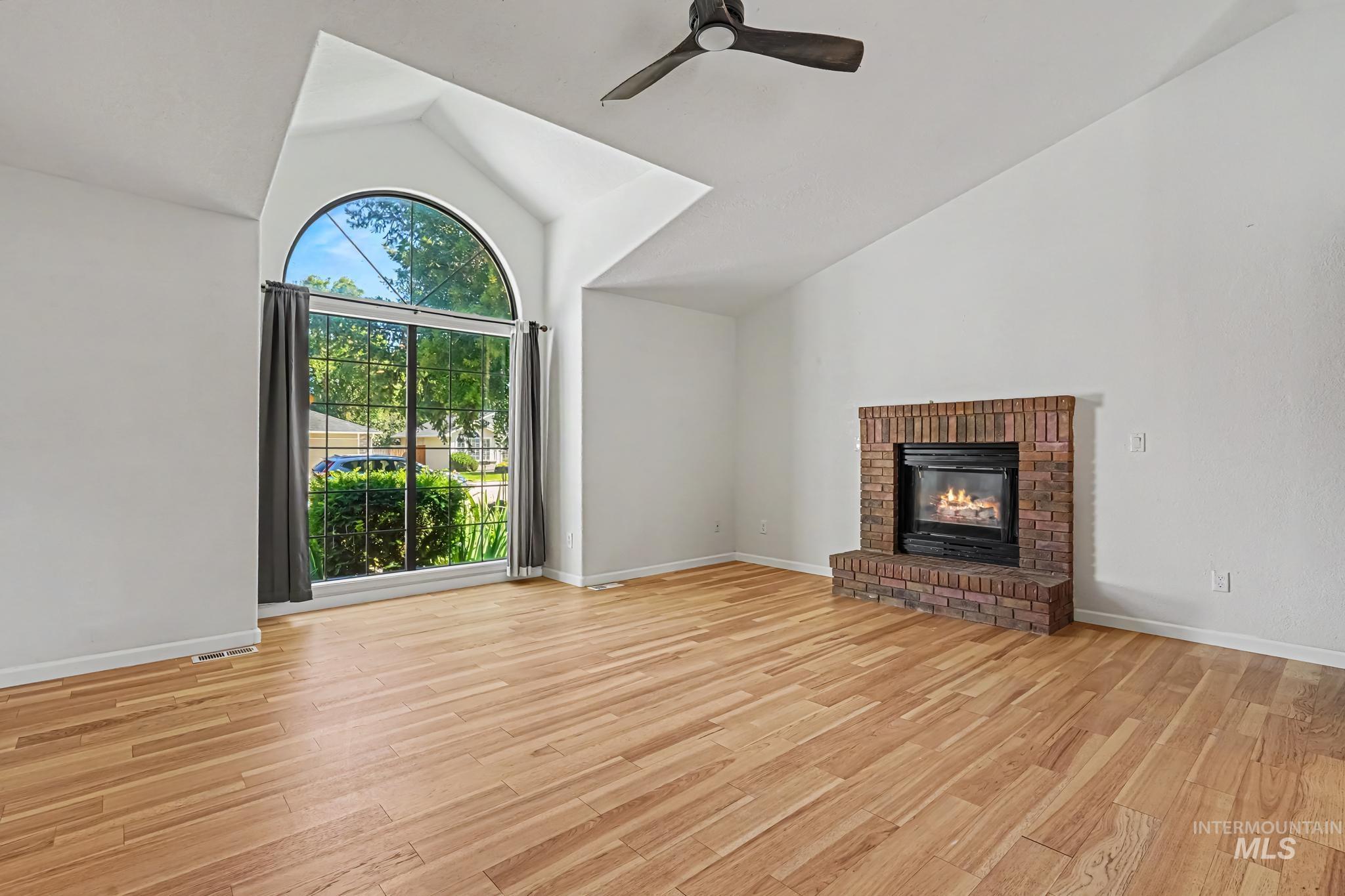 Spacious room featuring vaulted ceilings, a large arched window with exterior views, wood-finish flooring, a brick fireplace with a hearth, and a ceiling fan