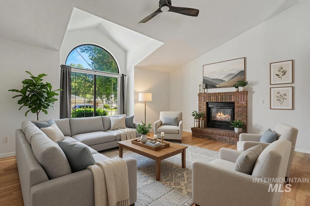 Spacious living room featuring vaulted ceilings, a large arched window, a brick-surround fireplace, a ceiling fan, and wood-finish flooring. *Virtually Staged photo*