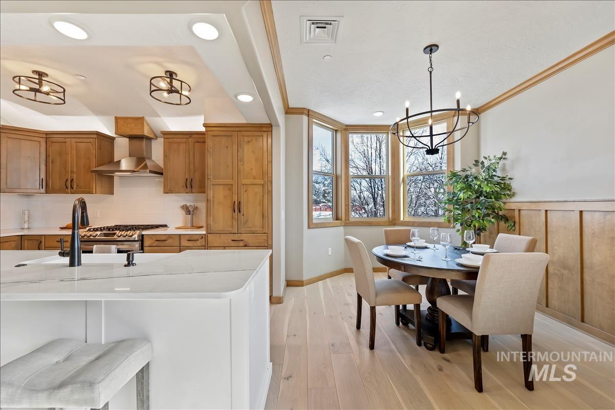 Kitchen featuring wall chimney exhaust hood, brown cabinetry, decorative backsplash, light wood-style flooring, and a chandelier