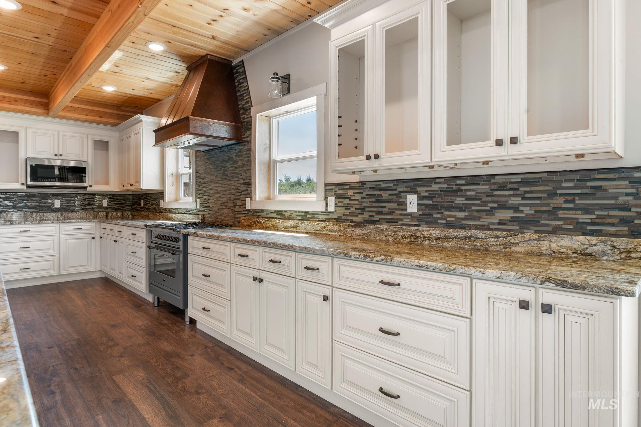Kitchen with dark wood-style flooring, custom range hood, white cabinetry, backsplash, and a wood ceiling with exposed beams