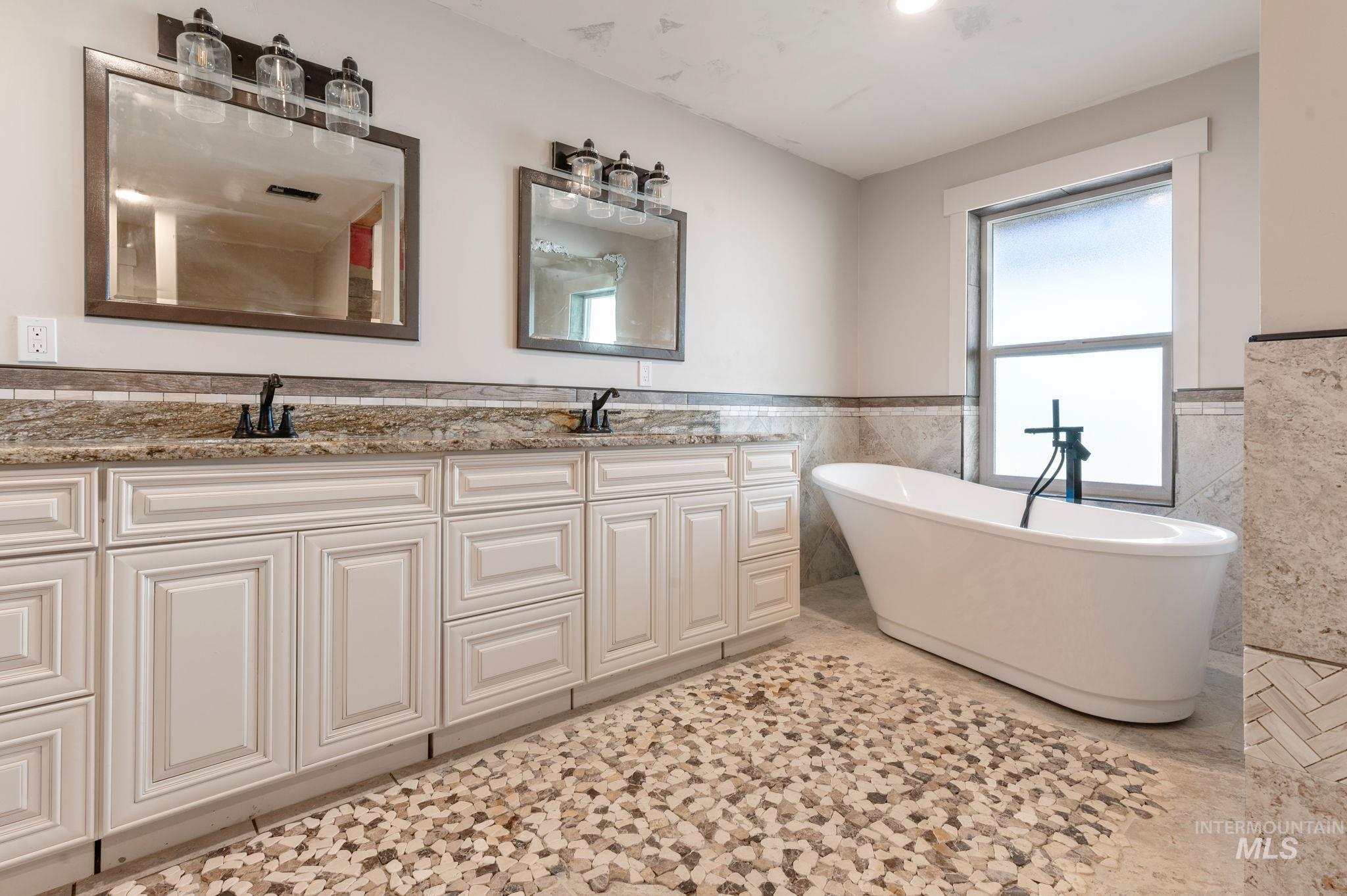 Bathroom featuring tile walls, a wainscoted wall, a freestanding tub, double vanity, and light tile patterned floors