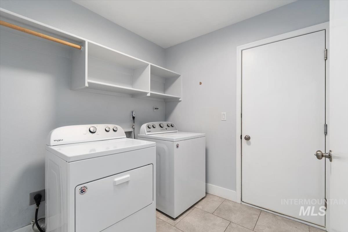 Dedicated laundry area featuring a white stackable washer and dryer, built-in open shelving, a wooden hanging rod, and light-toned tile flooring