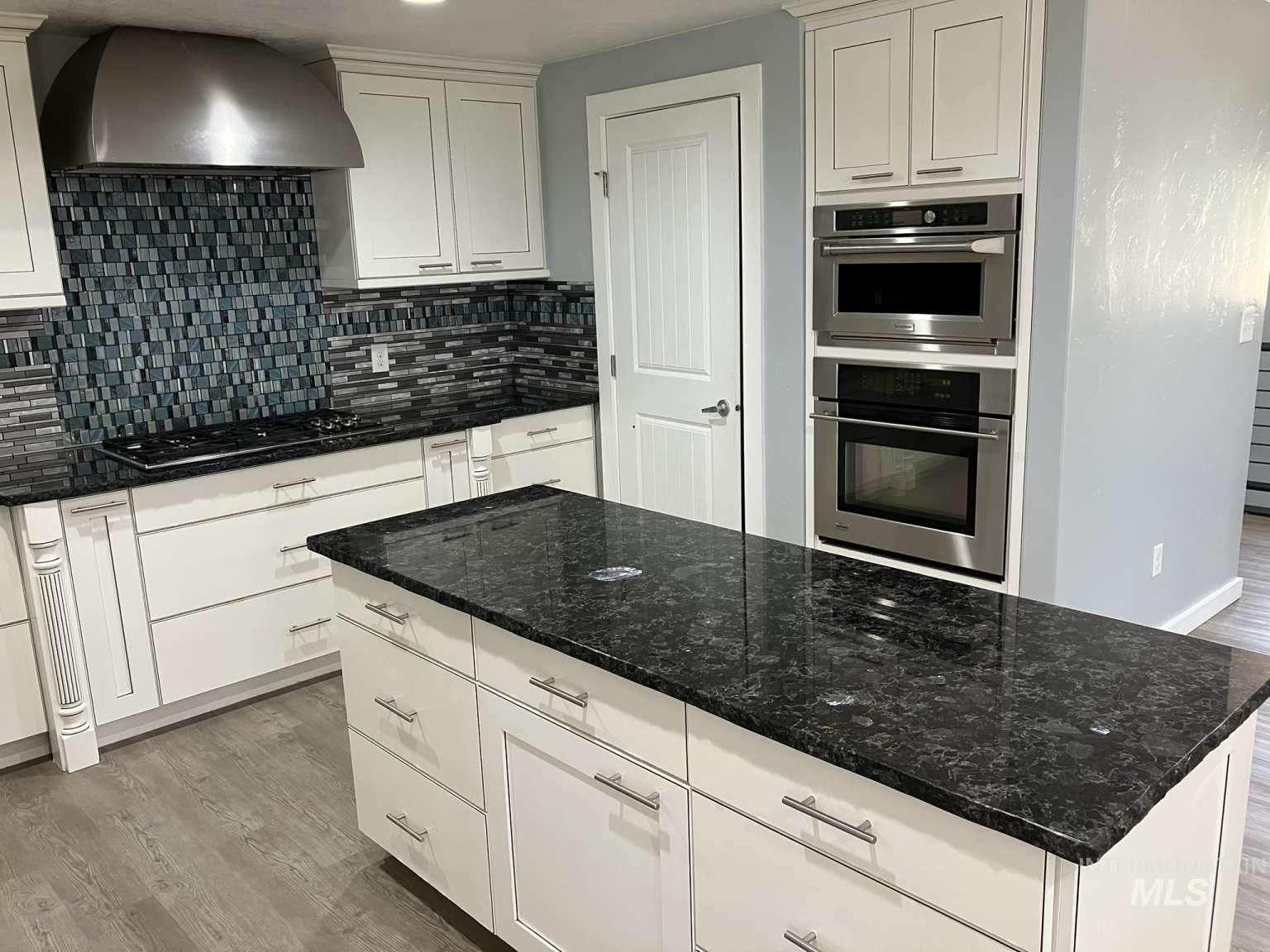 Kitchen with wall chimney exhaust hood, wood finished floors, backsplash, white cabinetry, and dark stone countertops