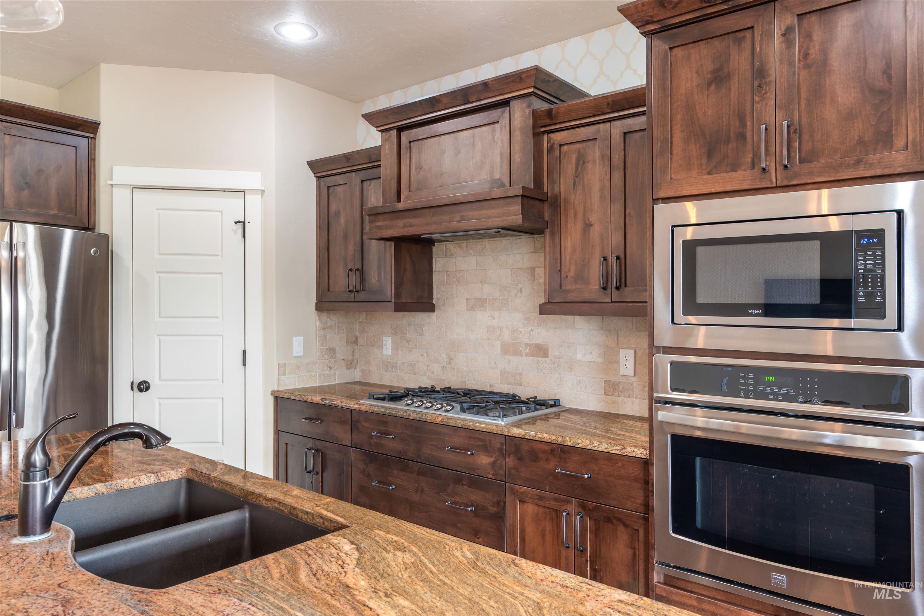 Kitchen with stainless steel appliances, light stone countertops, tasteful backsplash, premium range hood, and dark brown cabinets
