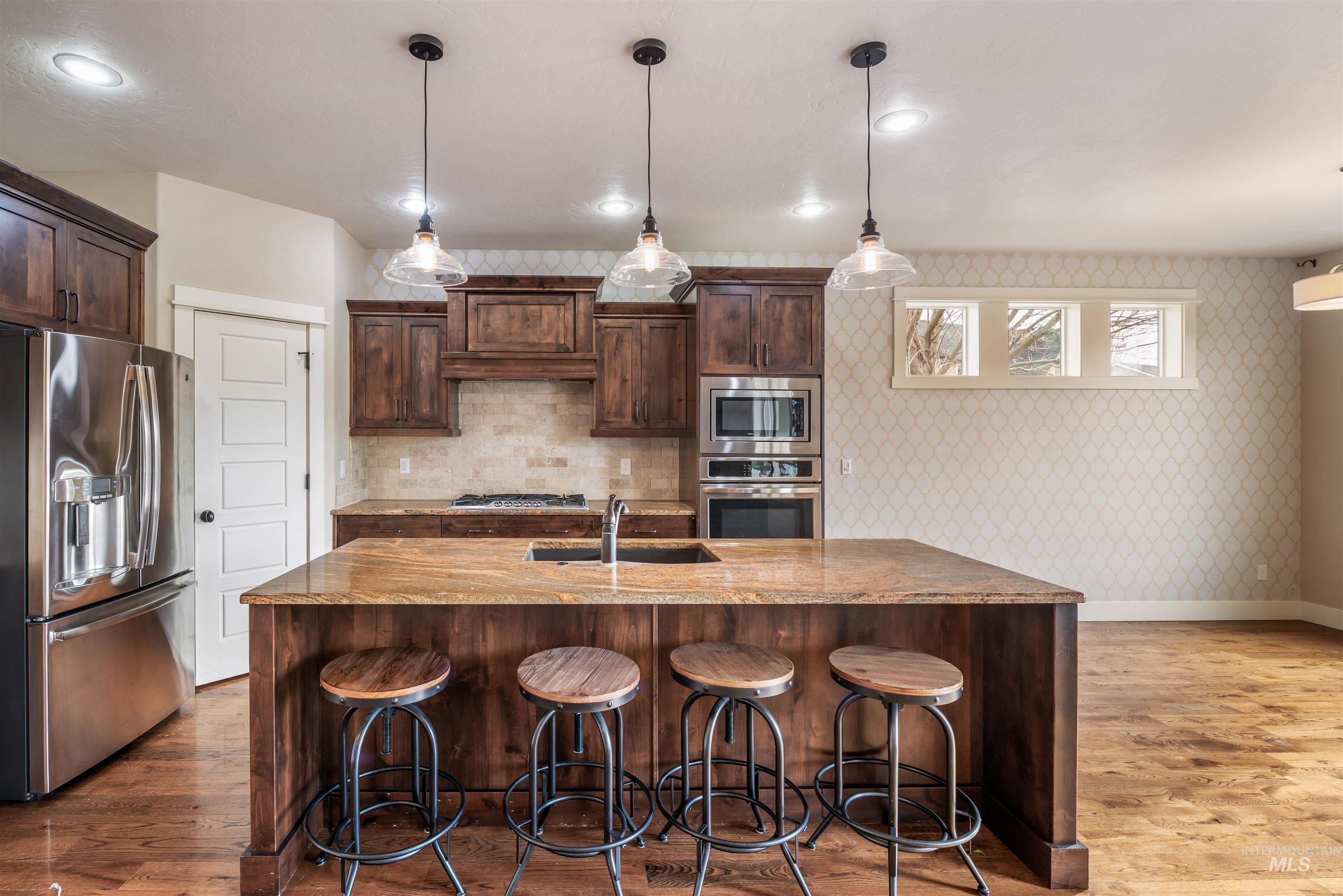 Kitchen with dark brown cabinetry, stainless steel appliances, hanging light fixtures, dark wood-type flooring, and a kitchen island with sink