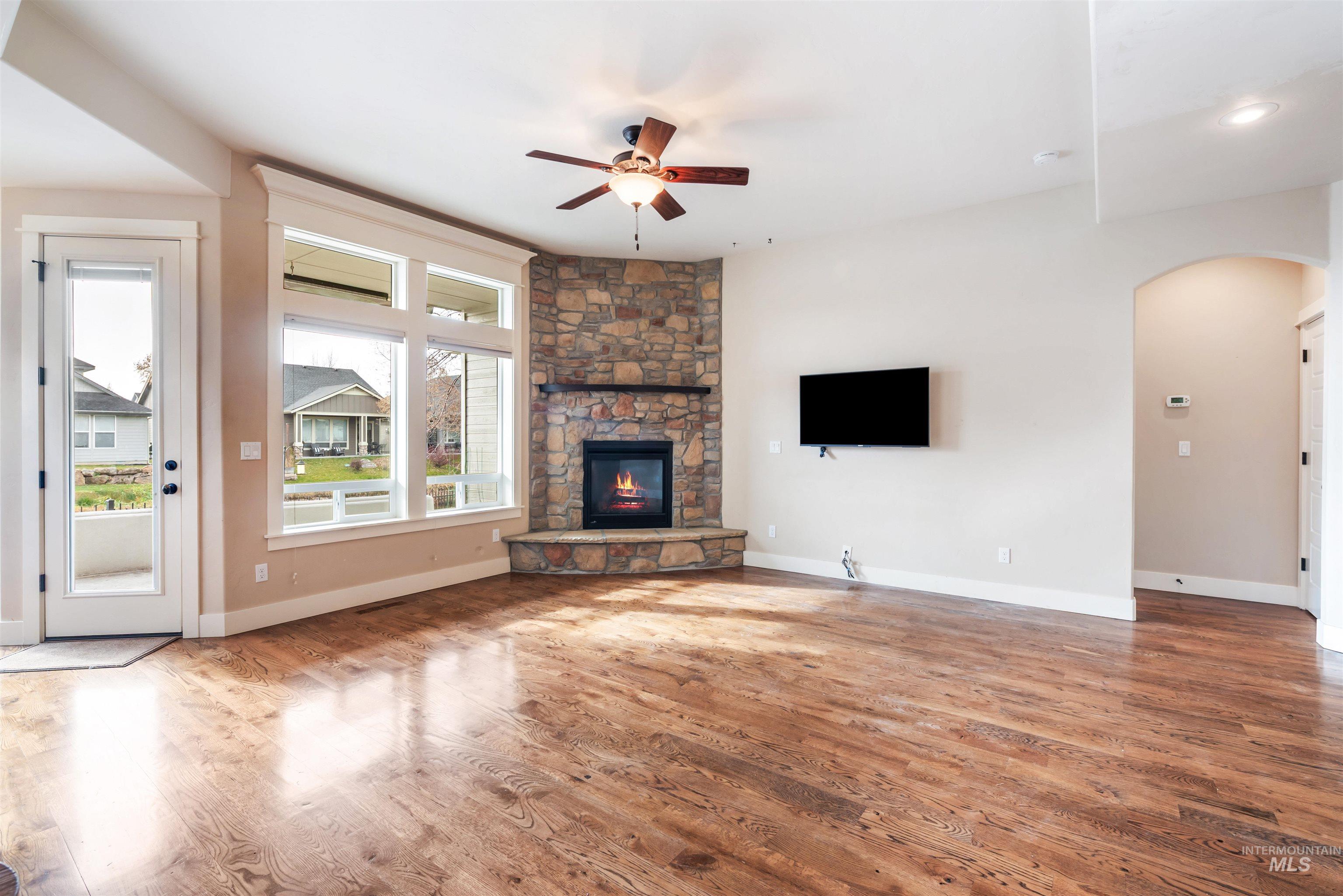Unfurnished living room featuring wood finished floors, arched walkways, ceiling fan, and a stone fireplace