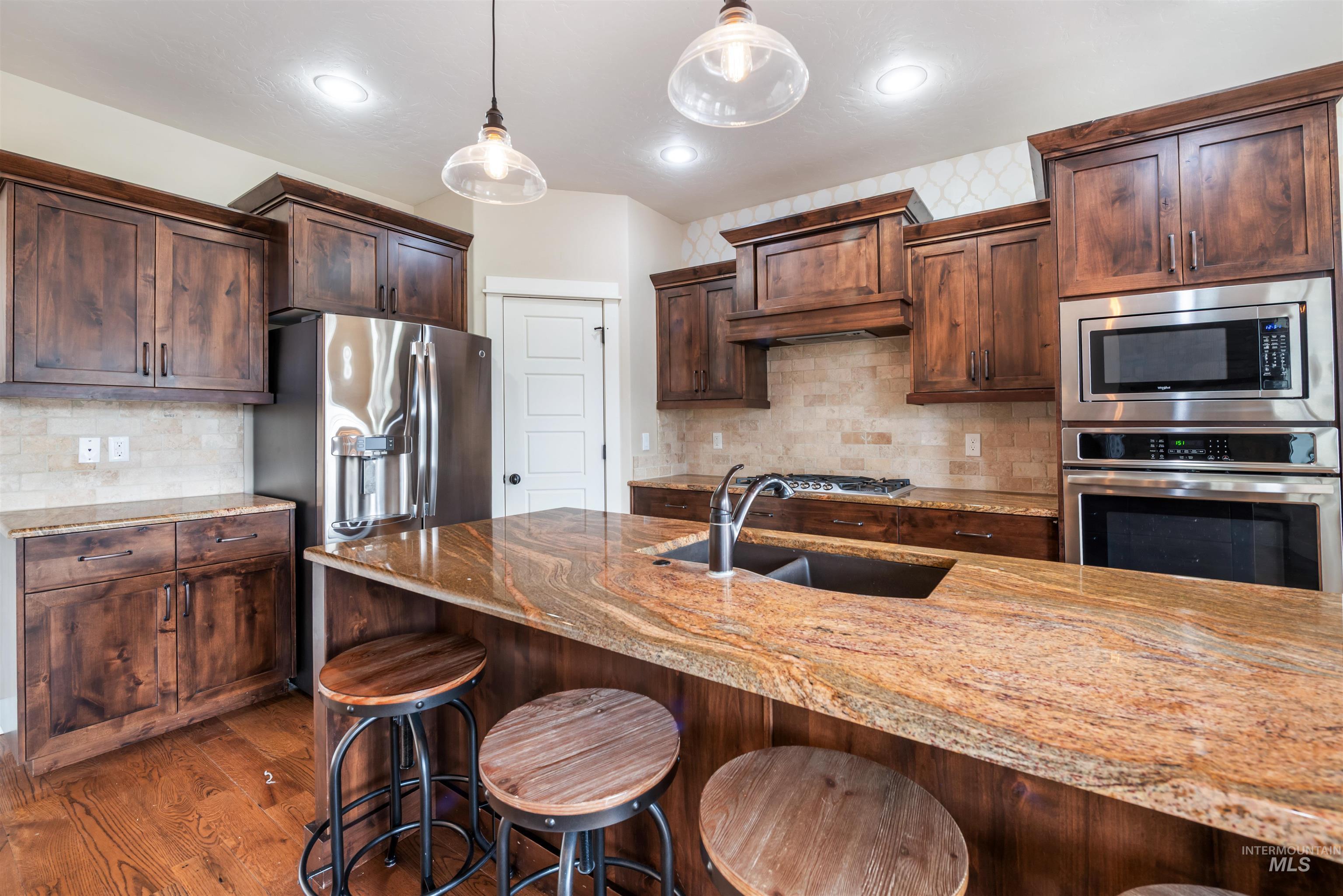 Kitchen with light stone counters, stainless steel appliances, hanging light fixtures, dark brown cabinets, and decorative backsplash