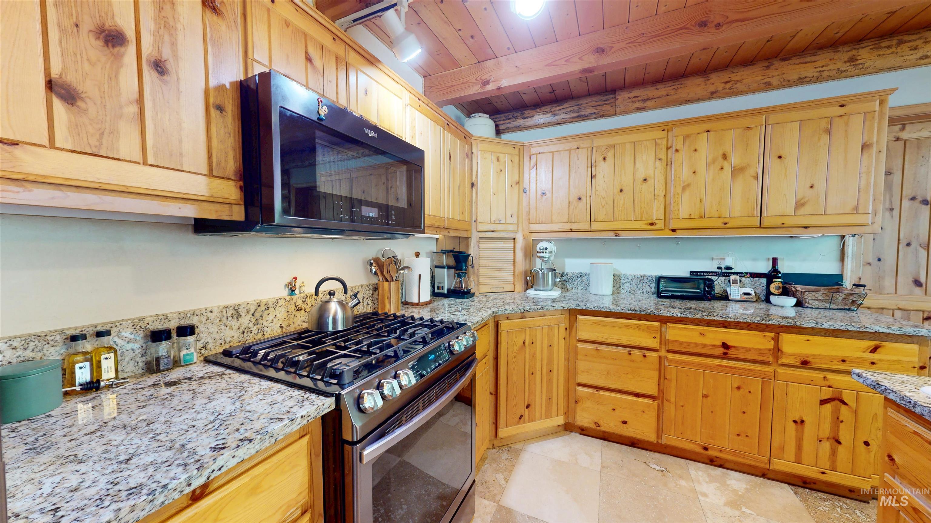 Kitchen with stainless steel range with gas stovetop, black microwave, a wooden ceiling with exposed beams, and light stone countertops