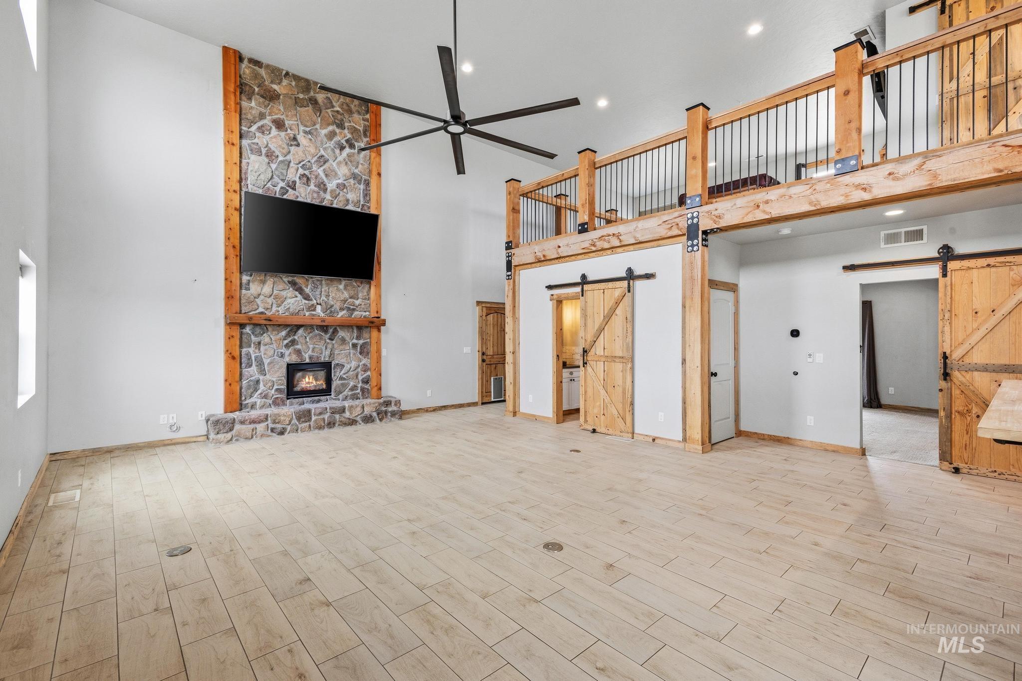 Two-story living area featuring a stone fireplace with a wood mantle, a large ceiling fan, wood-finish flooring, and a second-story loft with wood railing