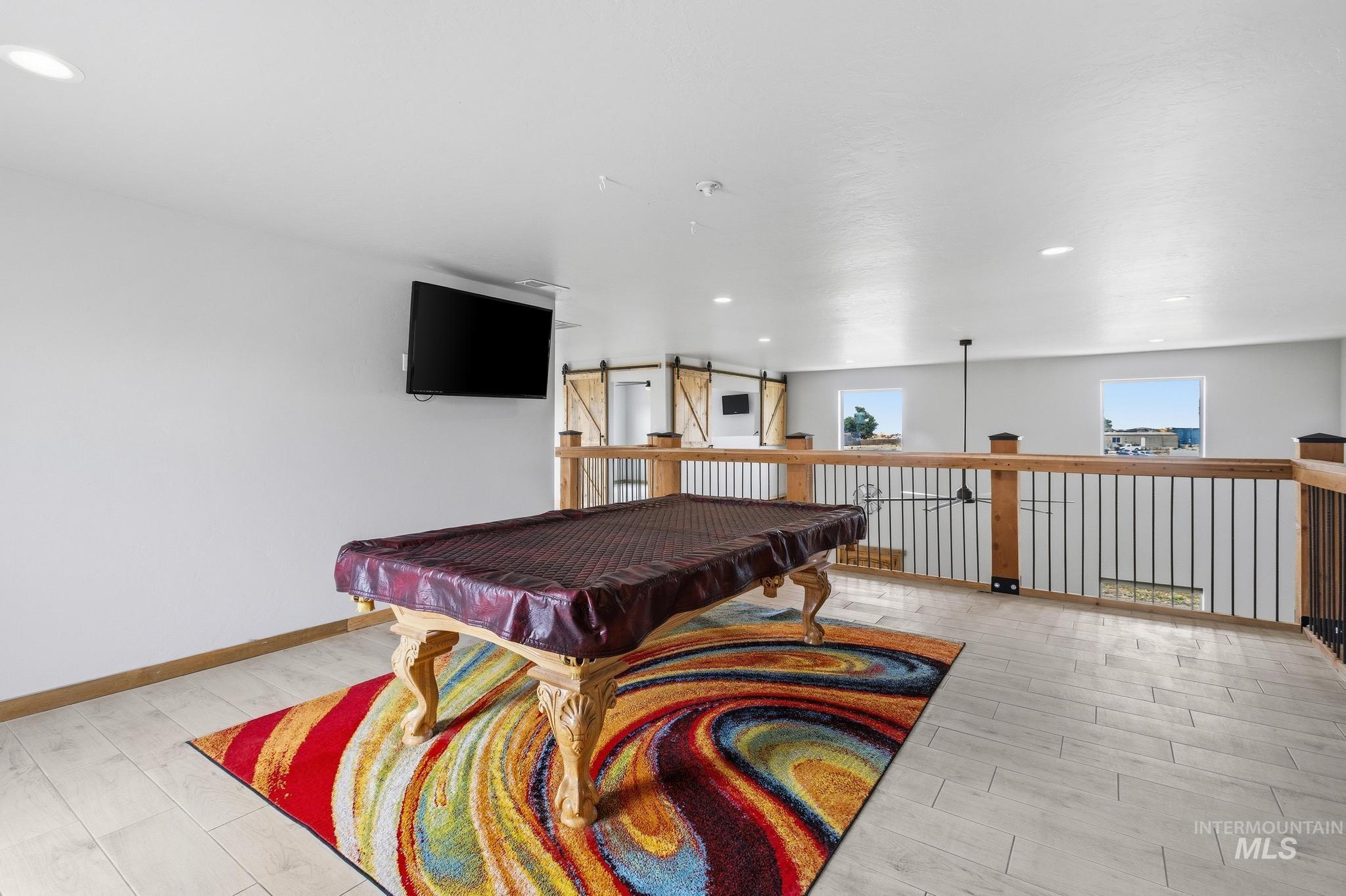Spacious loft area featuring light wood-finish flooring, a wall-mounted display, and a decorative railing system with wood accents and black balusters