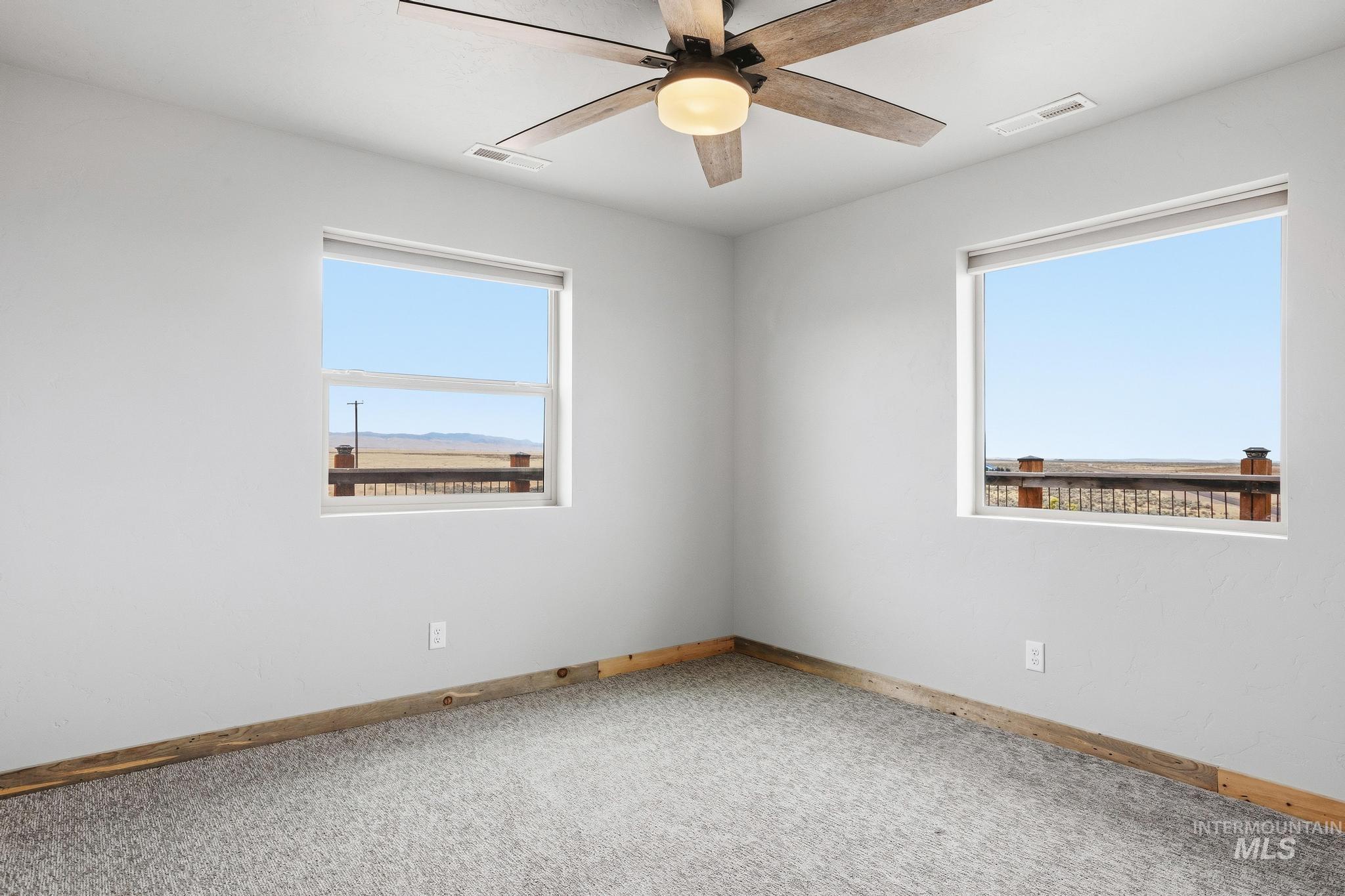 Bright room featuring two windows, a wood-finish ceiling fan with integrated lighting, light gray carpeting, and rustic wood base trim