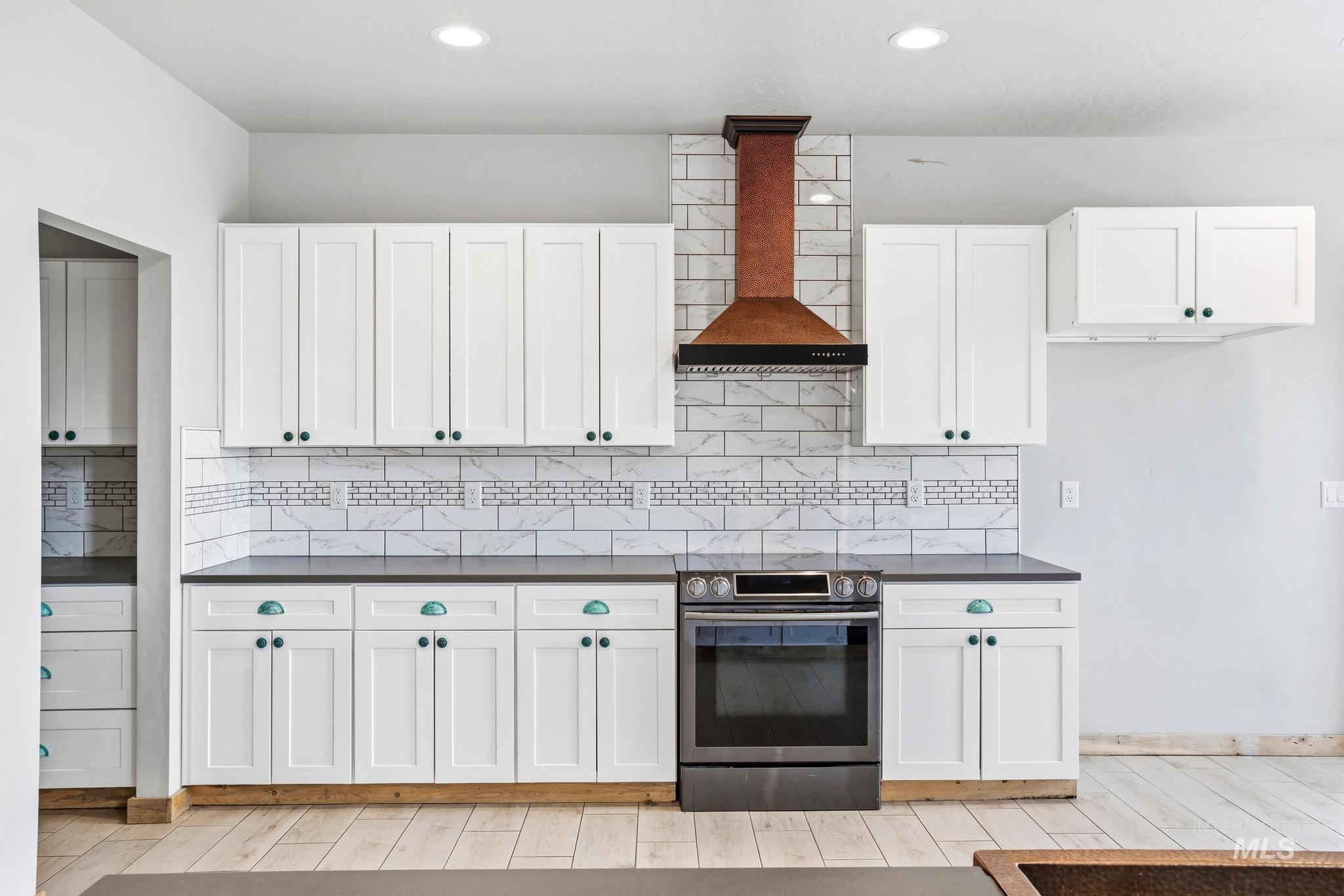 Kitchen featuring white shaker cabinetry, dark countertops, a tile backsplash with decorative mosaic inlay, a stainless steel range, and a copper-toned range hood